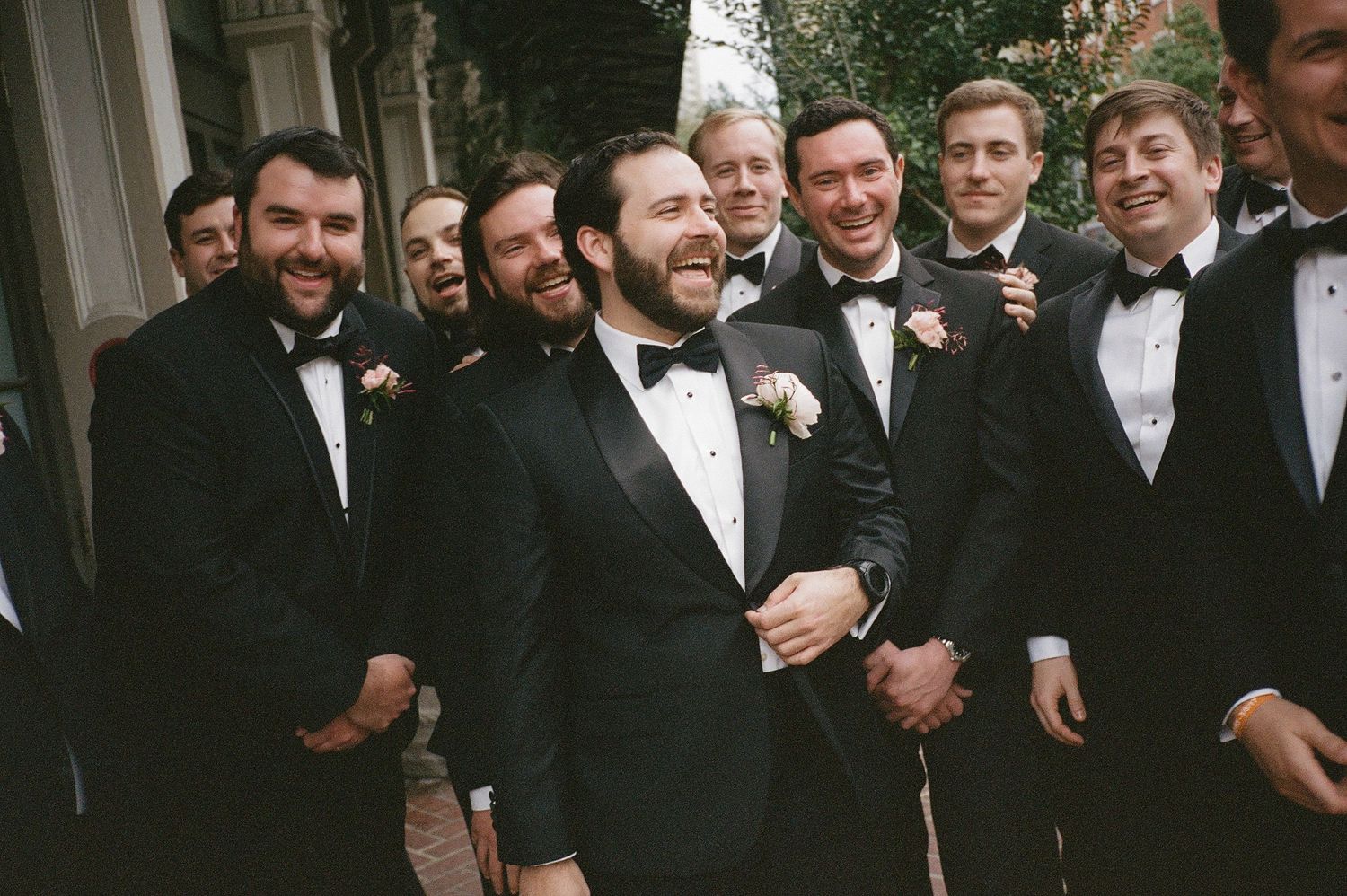 Groomsmen in black tuxedos laugh and celebrate together at a formal wedding event.
