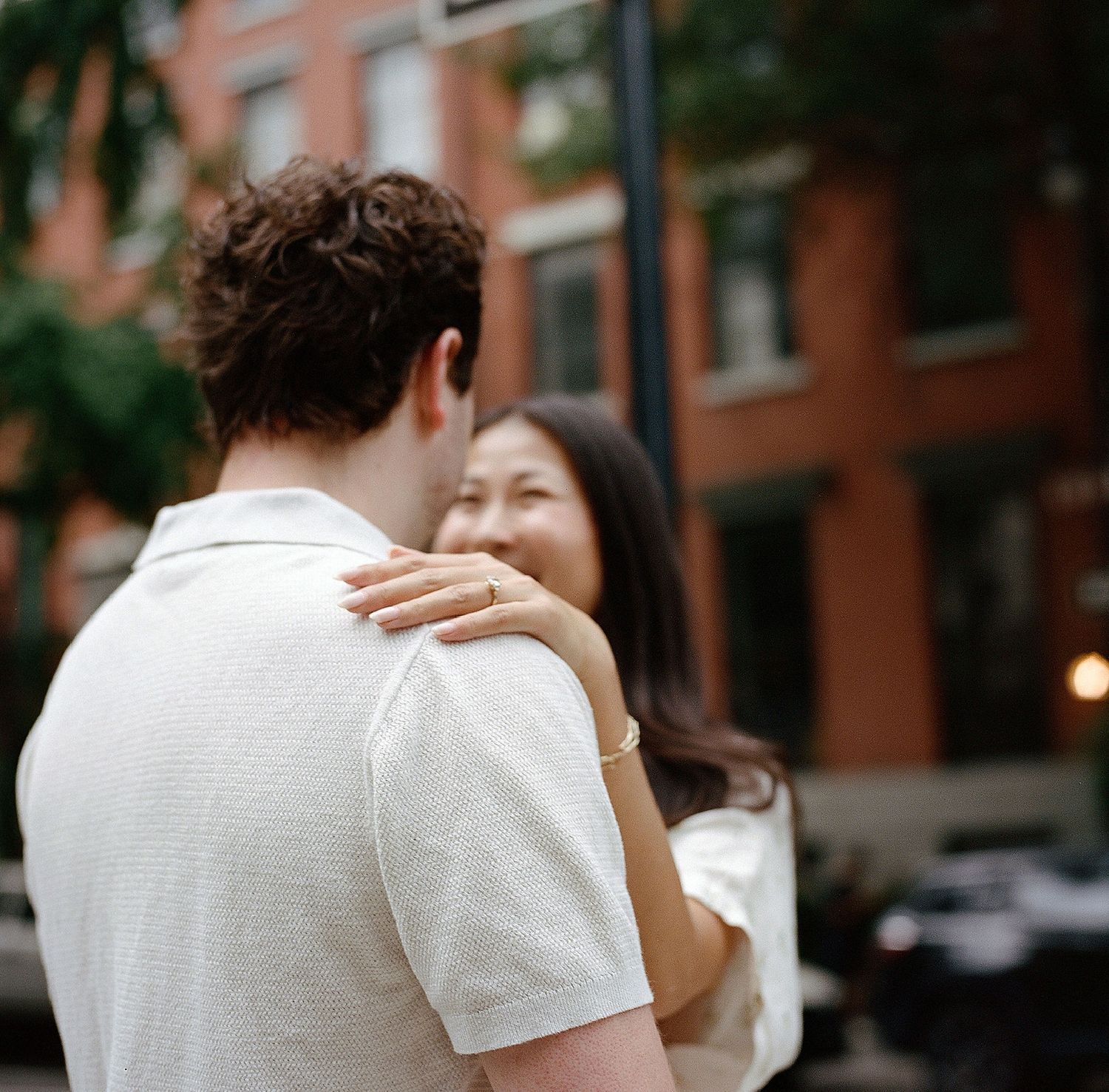 Couple embracing on a city street, woman smiling with hand on man's shoulder, urban brick building backdrop.