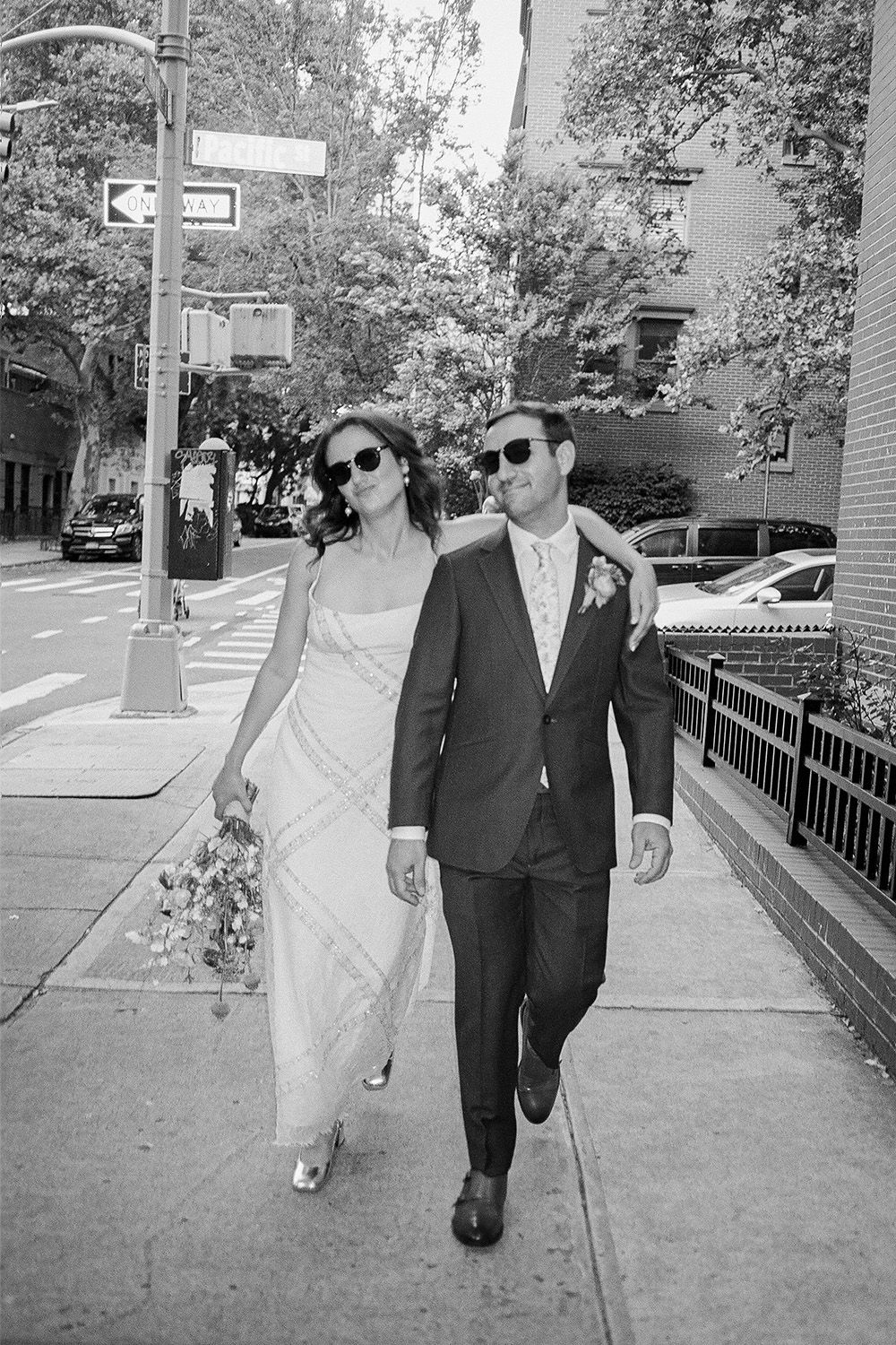 Wedding couple walking down city sidewalk wearing sunglasses.