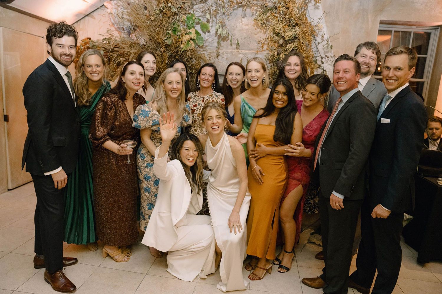 Wedding party group photo with guests in formal attire against an elegant neutral backdrop.