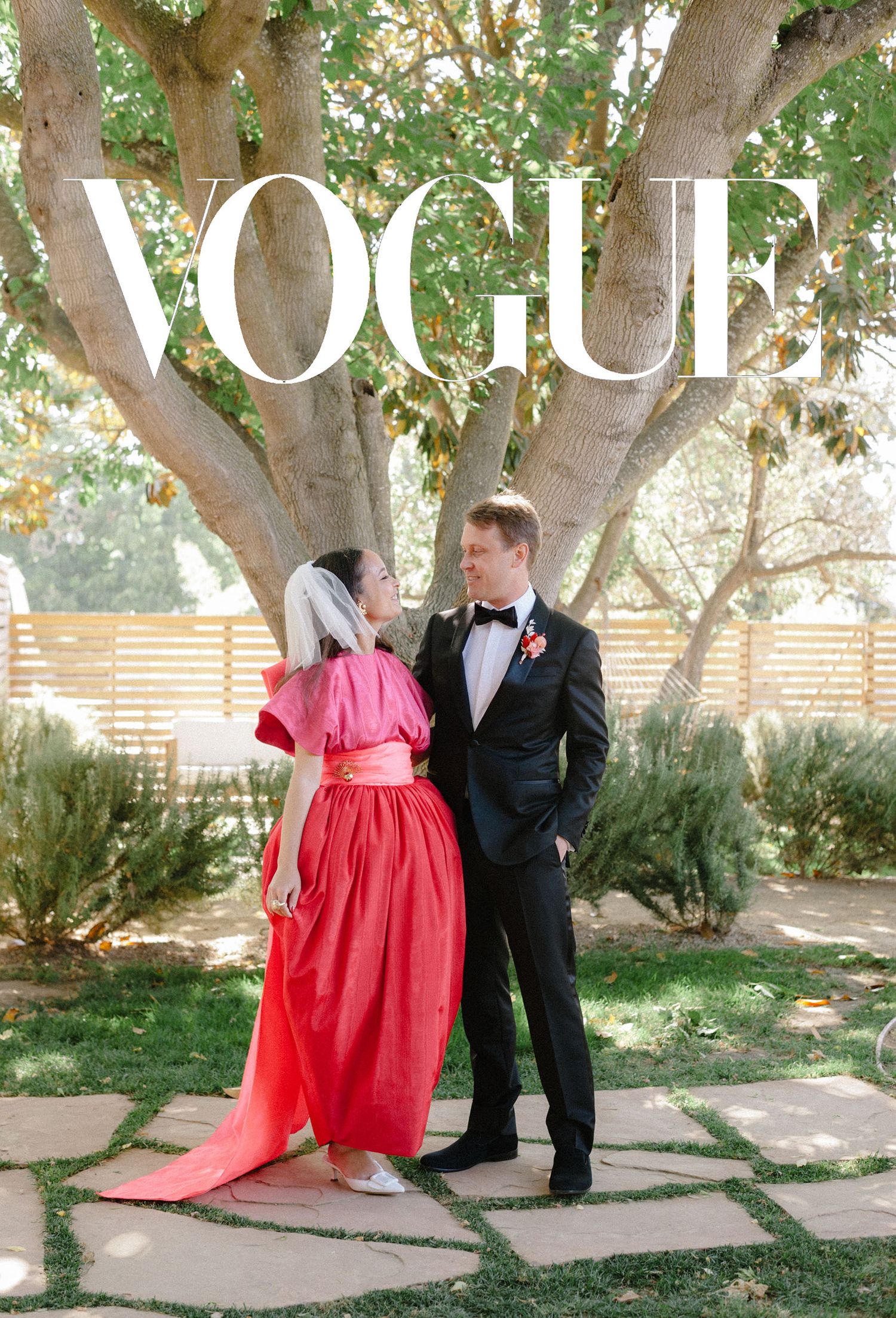 Wedding party members in formal attire and red dresses pose under shady trees in a garden.
