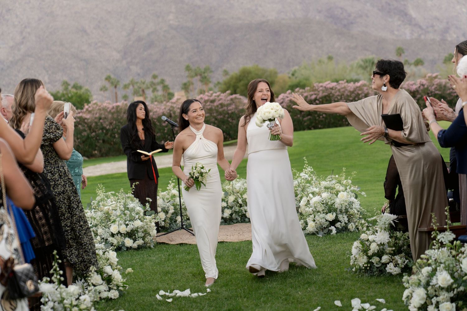 Wedding celebration on a lawn with floral decorations and mountain backdrop as guests cheer.