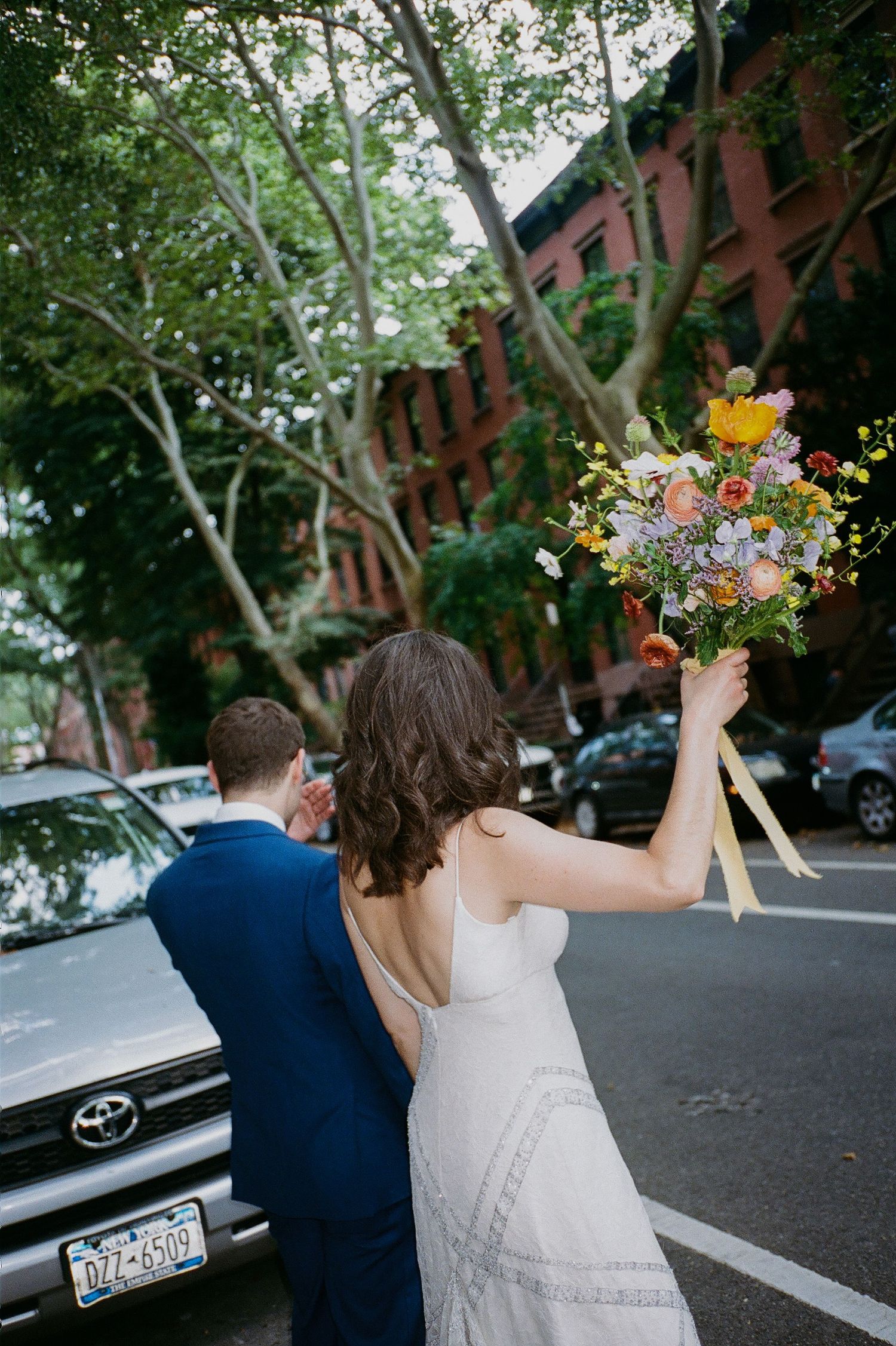 Bride in white dress raises colorful bouquet while walking with groom in blue suit on a New York City street.