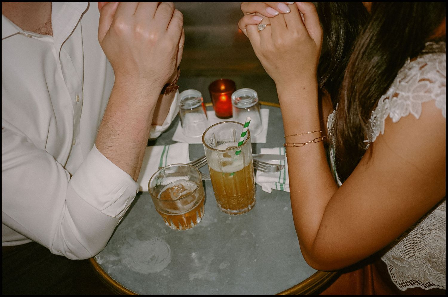 Two people sit at a candlelit bar table with drinks, their faces hidden as they lean forward together.