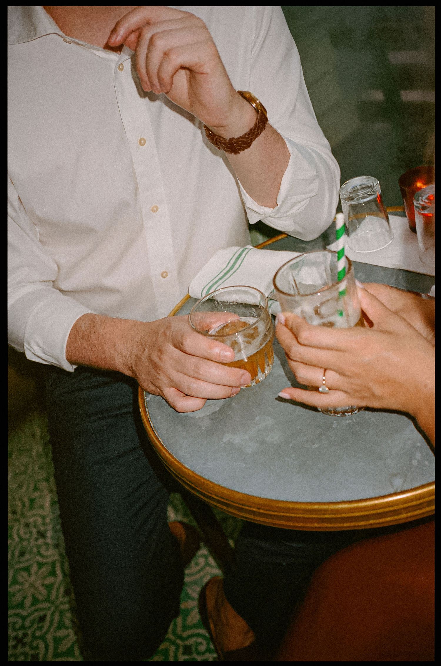 Two people clinking glasses at a small round bar table, one wearing a gold watch and the other a diamond ring.