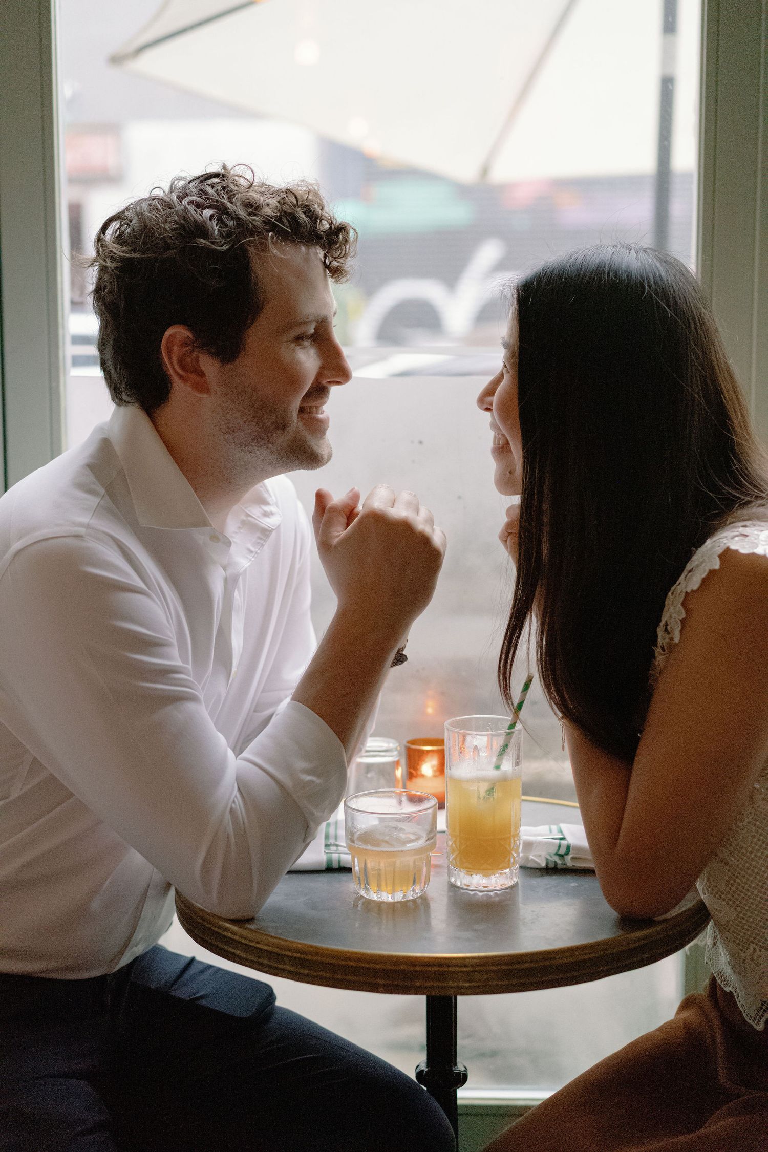 Couple holding hands at a romantic cafe table with candles and drinks, sharing an intimate moment by a window.