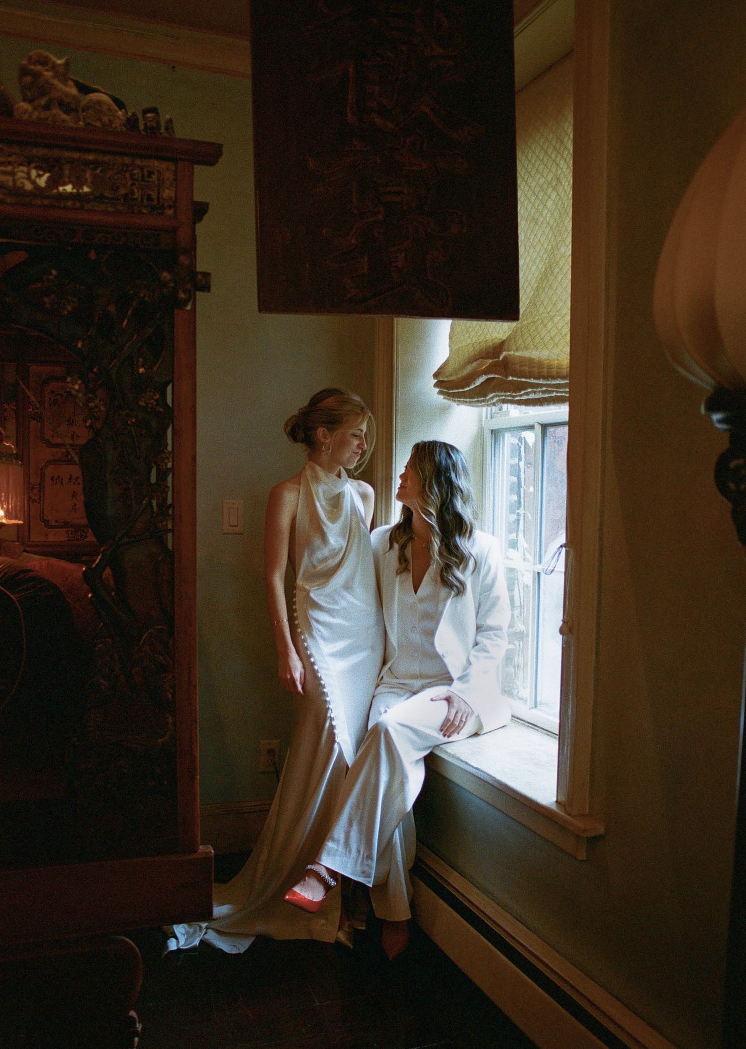 Two figures in white dresses stand by a window in an ornate room with vintage wooden furniture.