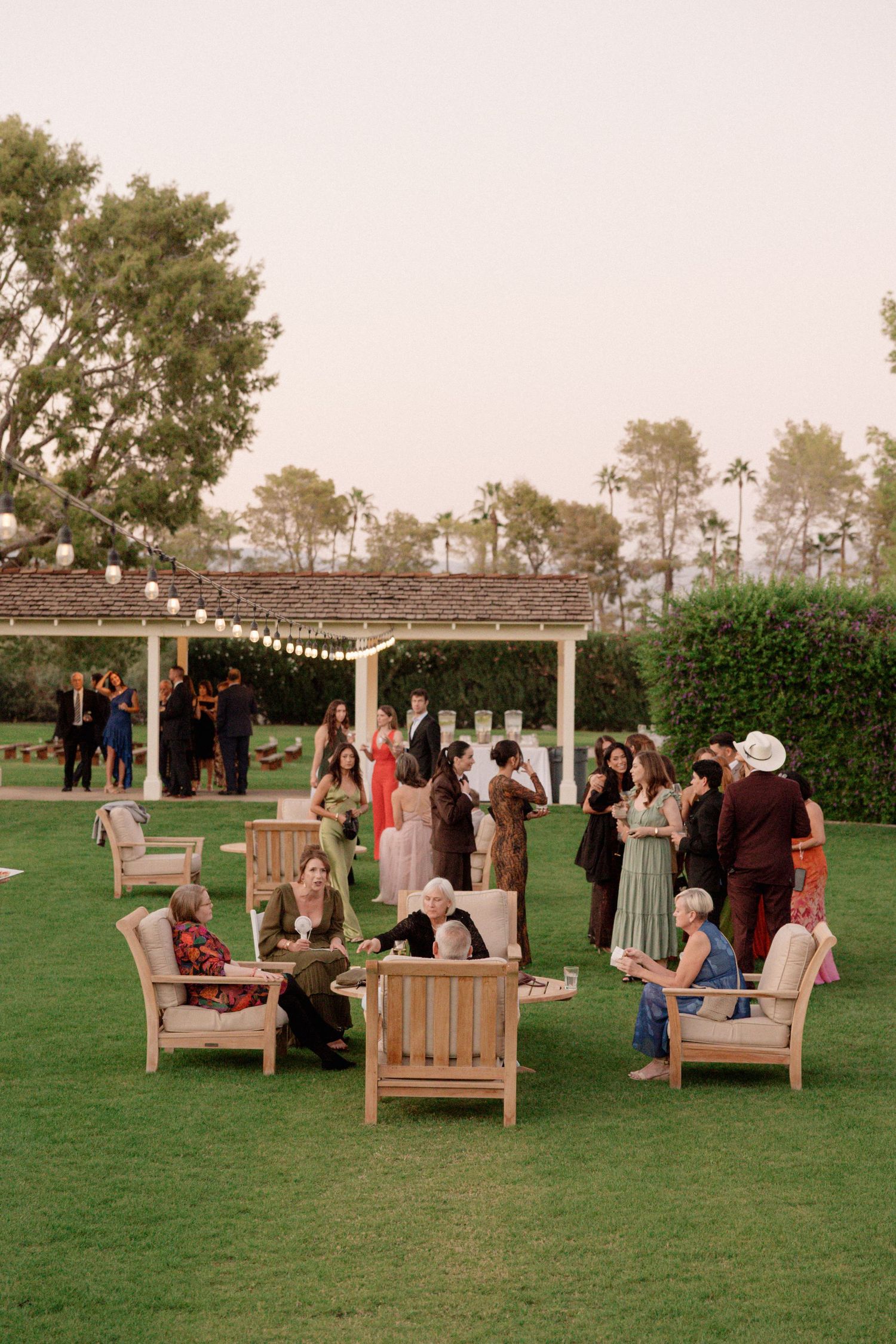 Outdoor wedding reception seating with wooden benches arranged on a green lawn under string lights at dusk.