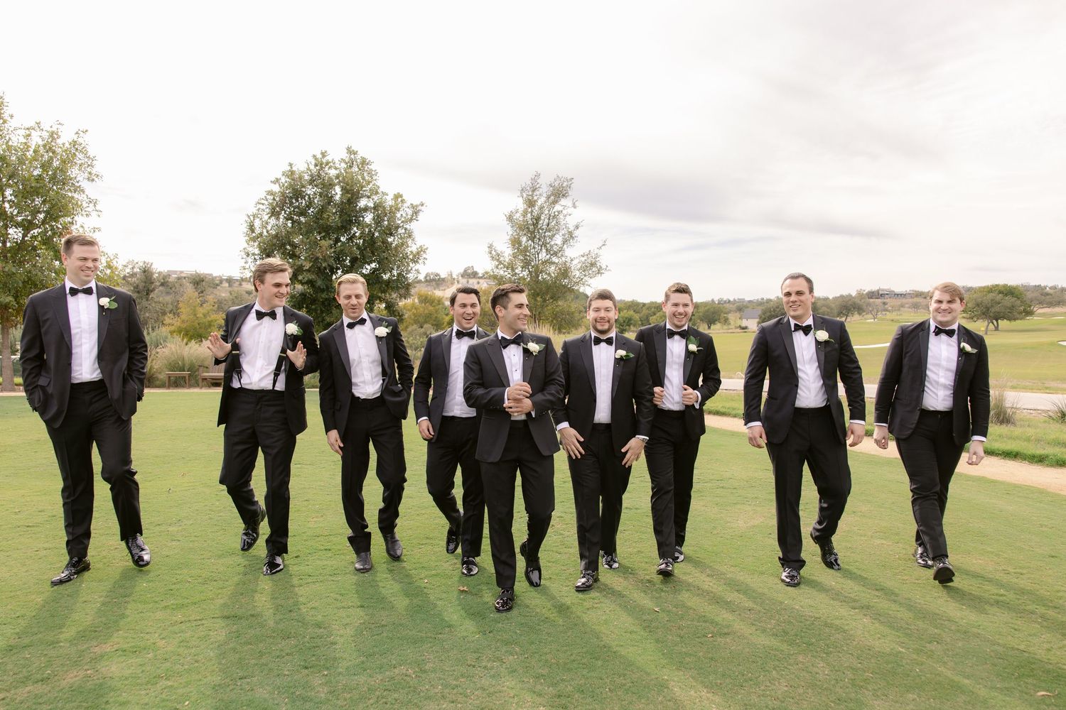 Groomsmen in black tuxedos lined up on a green lawn for a wedding photo.