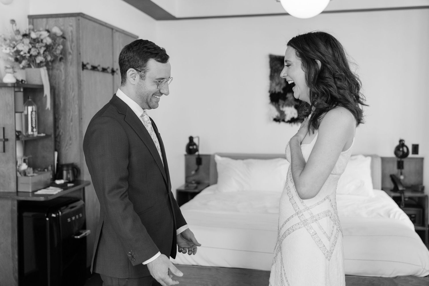 Black and white photo of a couple sharing a joyful moment in a hotel room, man in suit, woman in white dress.