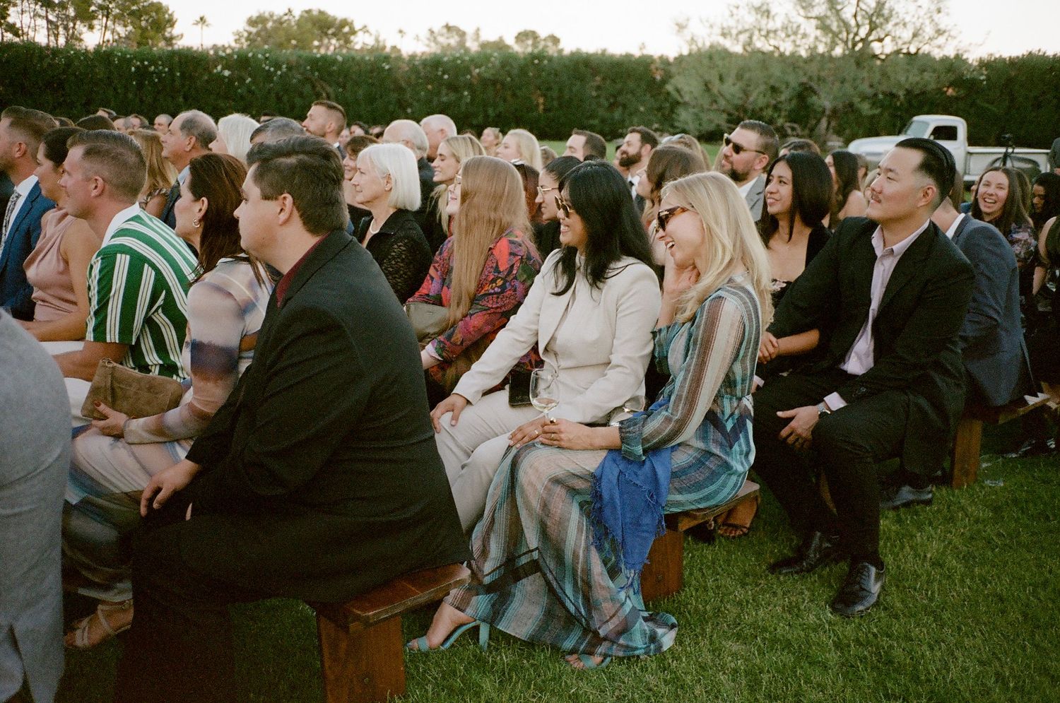 Wedding guests seated on lawn chairs during outdoor ceremony with green hedges in background.
