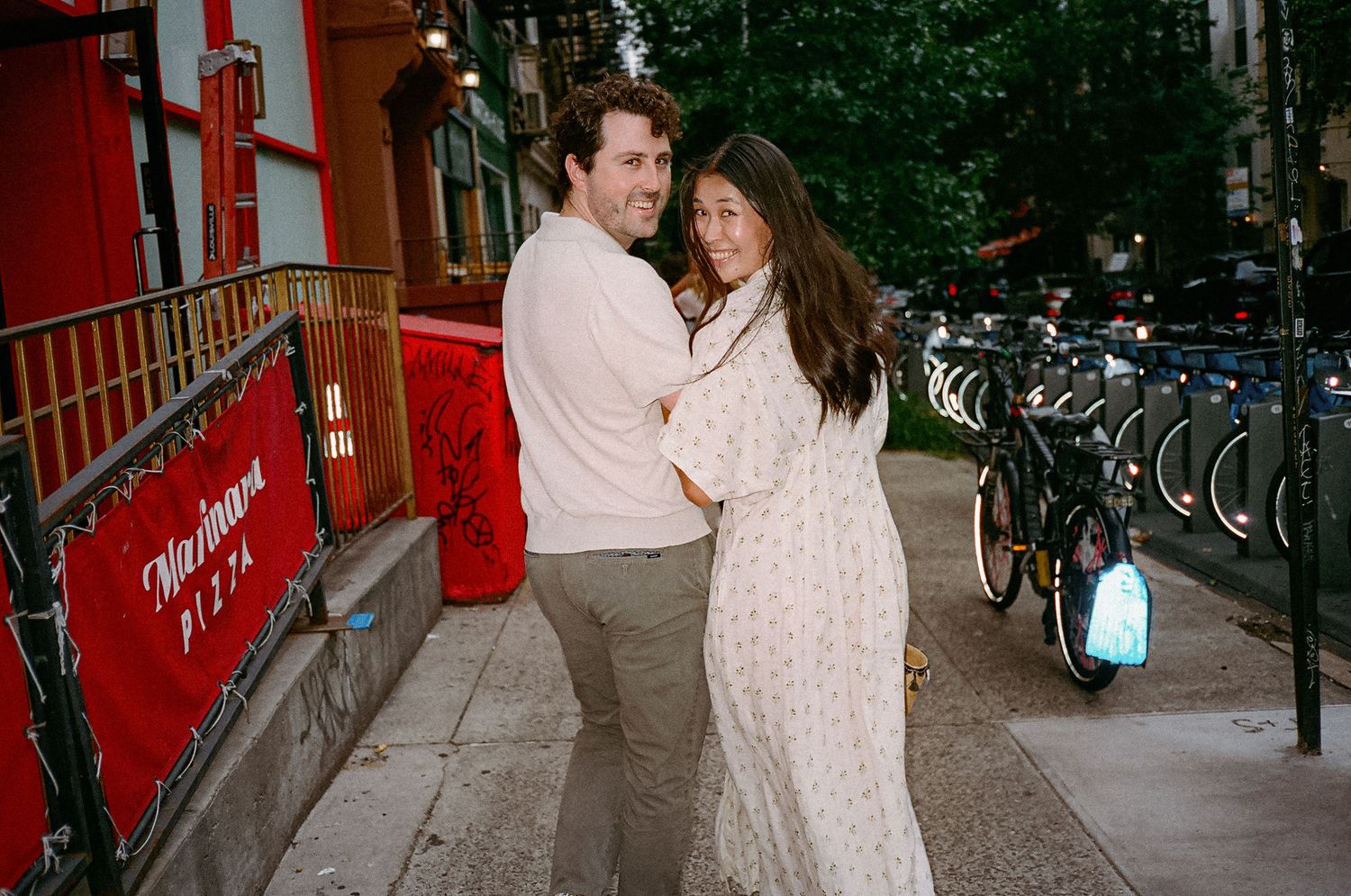 Couple embracing and smiling on a city sidewalk near a pizza shop with bicycles in the background.