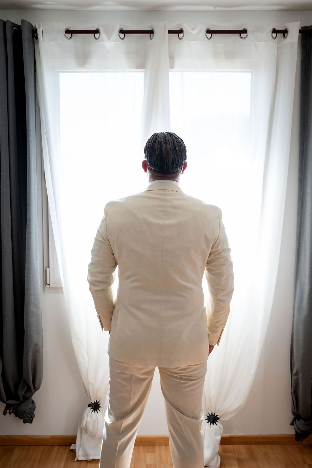 Man in white suit standing with back to camera gazing out bright window with gray and white curtains.
