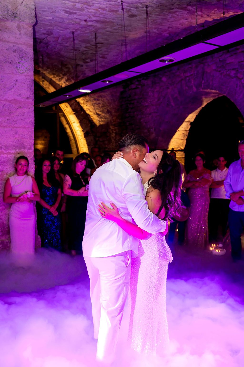 Bride and groom share first dance surrounded by fog and purple lights in a stone venue with guests watching.