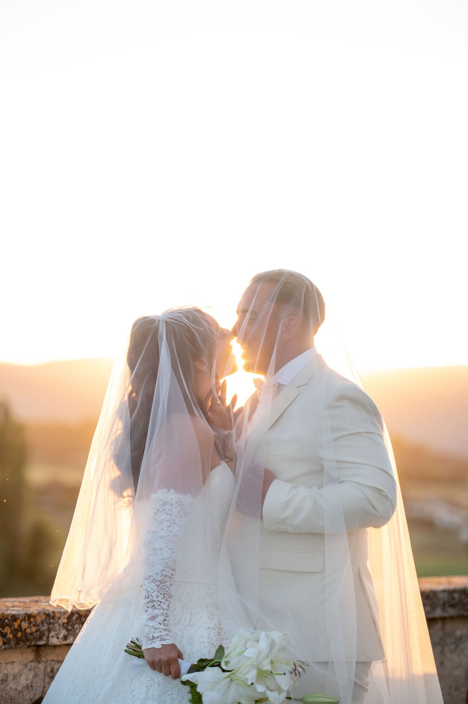 Bride and groom sharing a kiss under a veil at golden sunset, bride holding white lilies bouquet.
