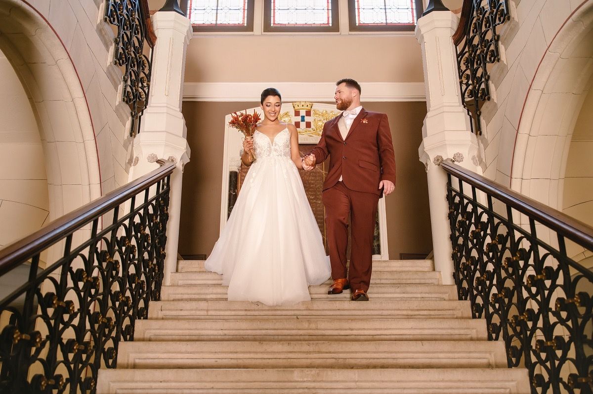 Wedding couple poses on elegant marble staircase with ornate black iron railings in classic architectural setting.