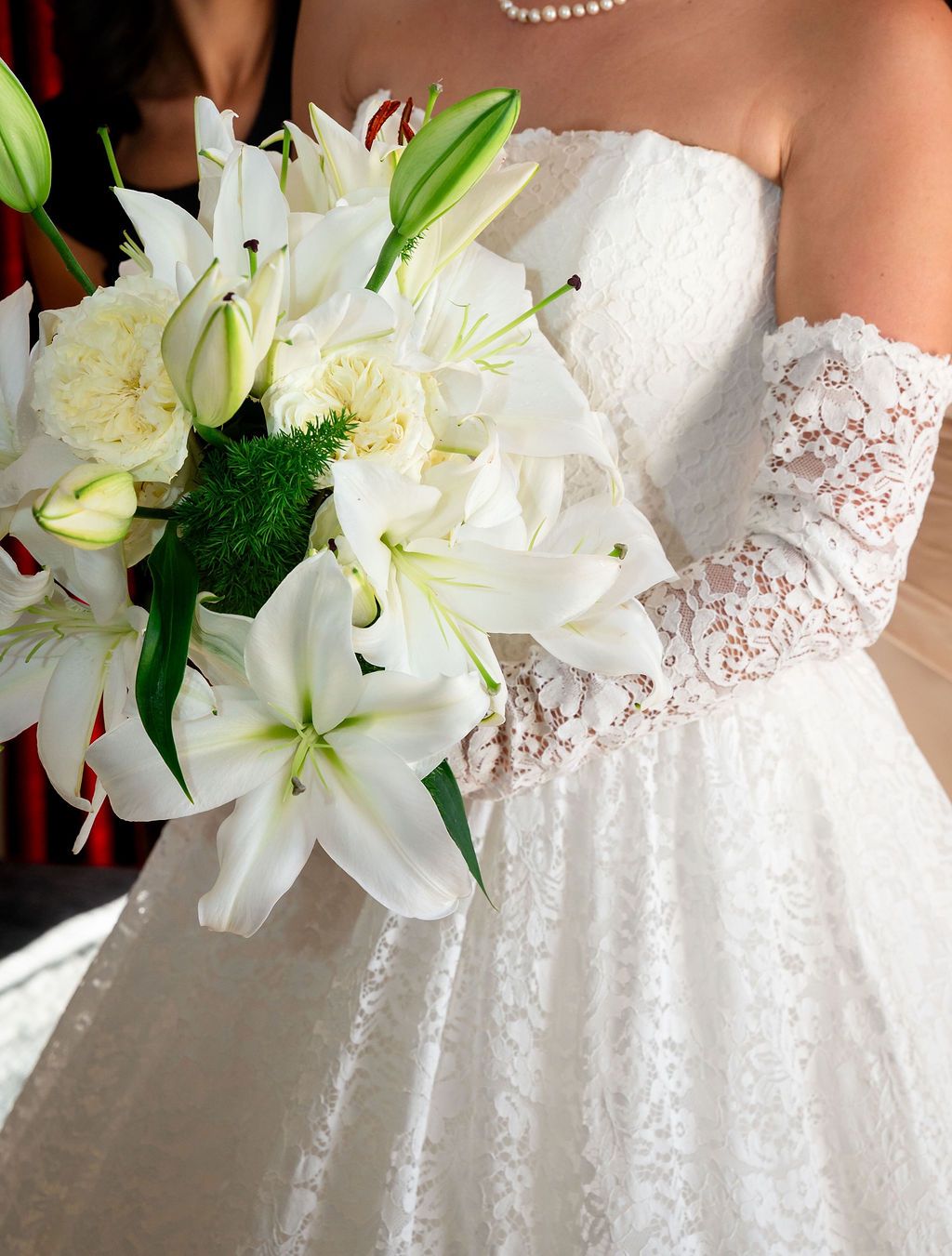 Bride in white lace gown and gloves holding elegant bouquet of white lilies and green accents on wedding day.
