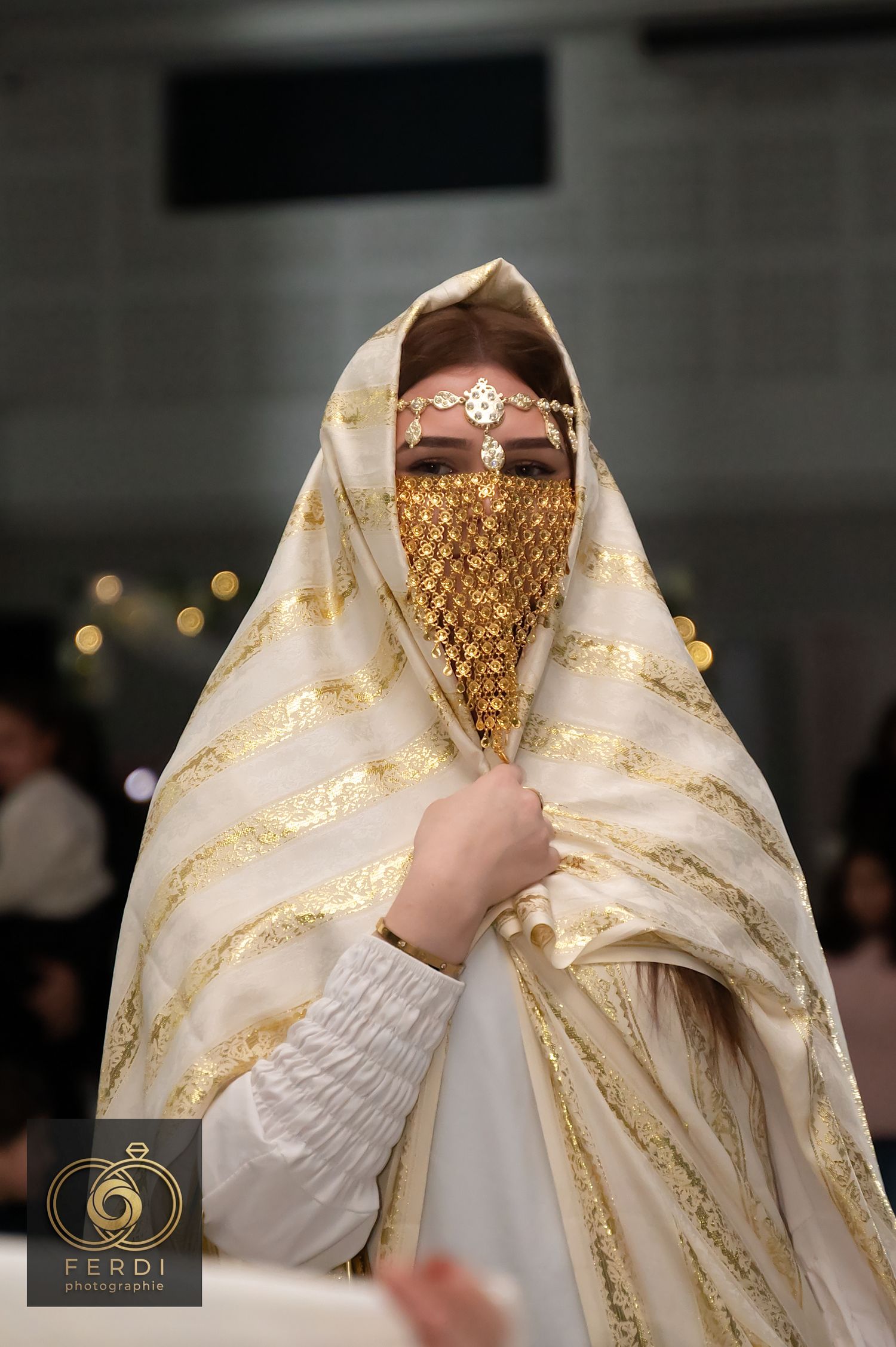 A bride wearing a traditional white and gold embroidered veil with an ornate gold face covering at a wedding ceremony.