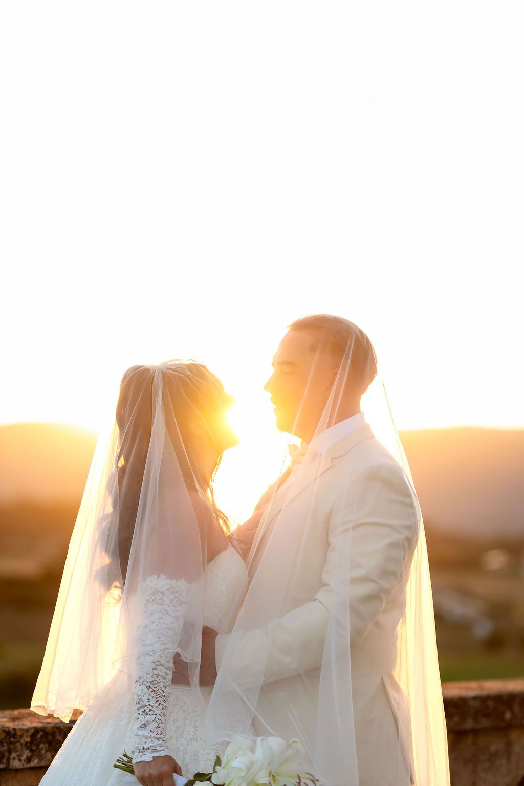 Bride and groom share a romantic sunset moment, bride in lace gown with veil, groom in white suit, holding flowers.