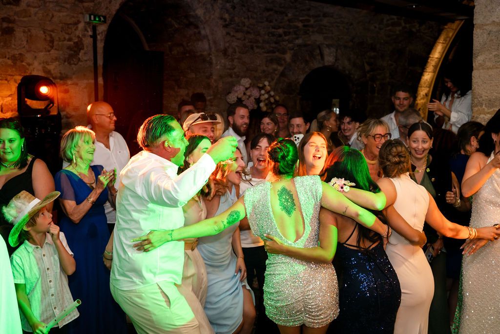 Guests dancing joyfully at a wedding reception under green lights in a rustic stone venue.