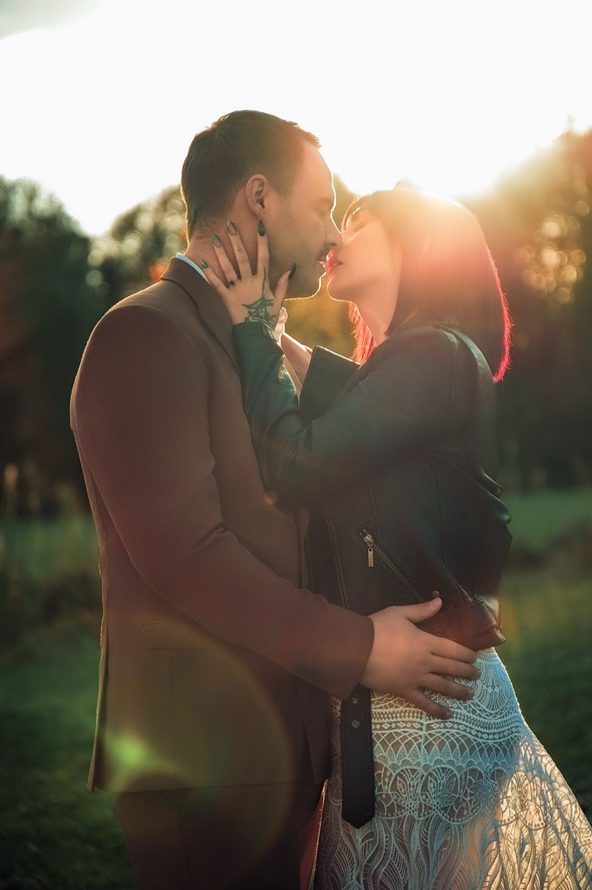 A romantic couple embraces and kisses in a sunlit field during golden hour with sun flare in the background.