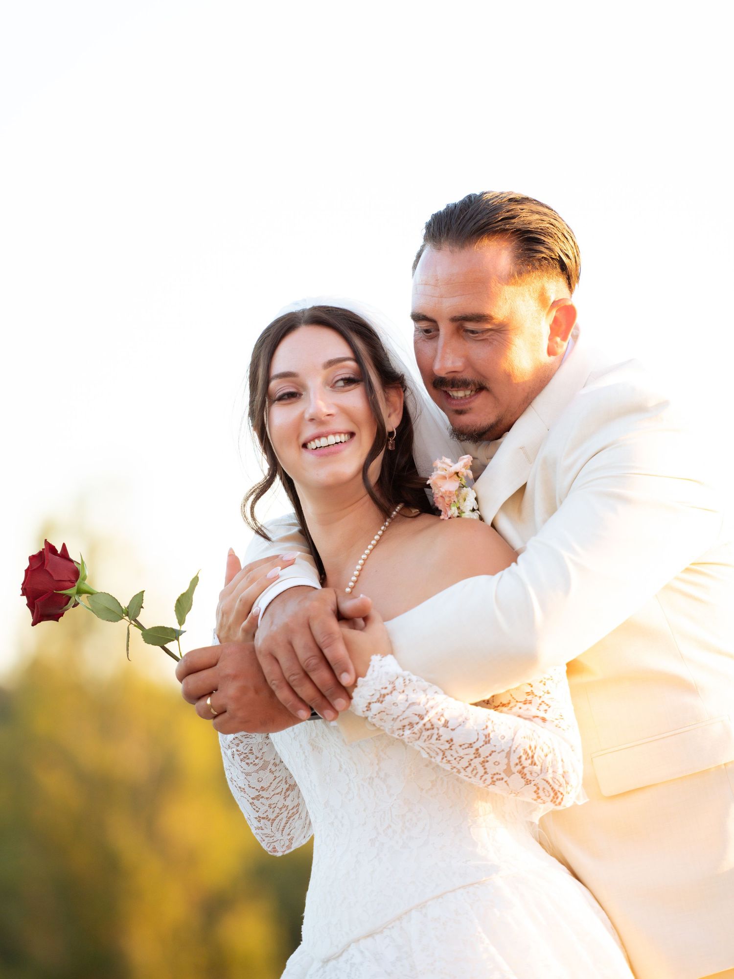 Bride in lace dress holds red rose while groom in white suit embraces her from behind outdoors.
