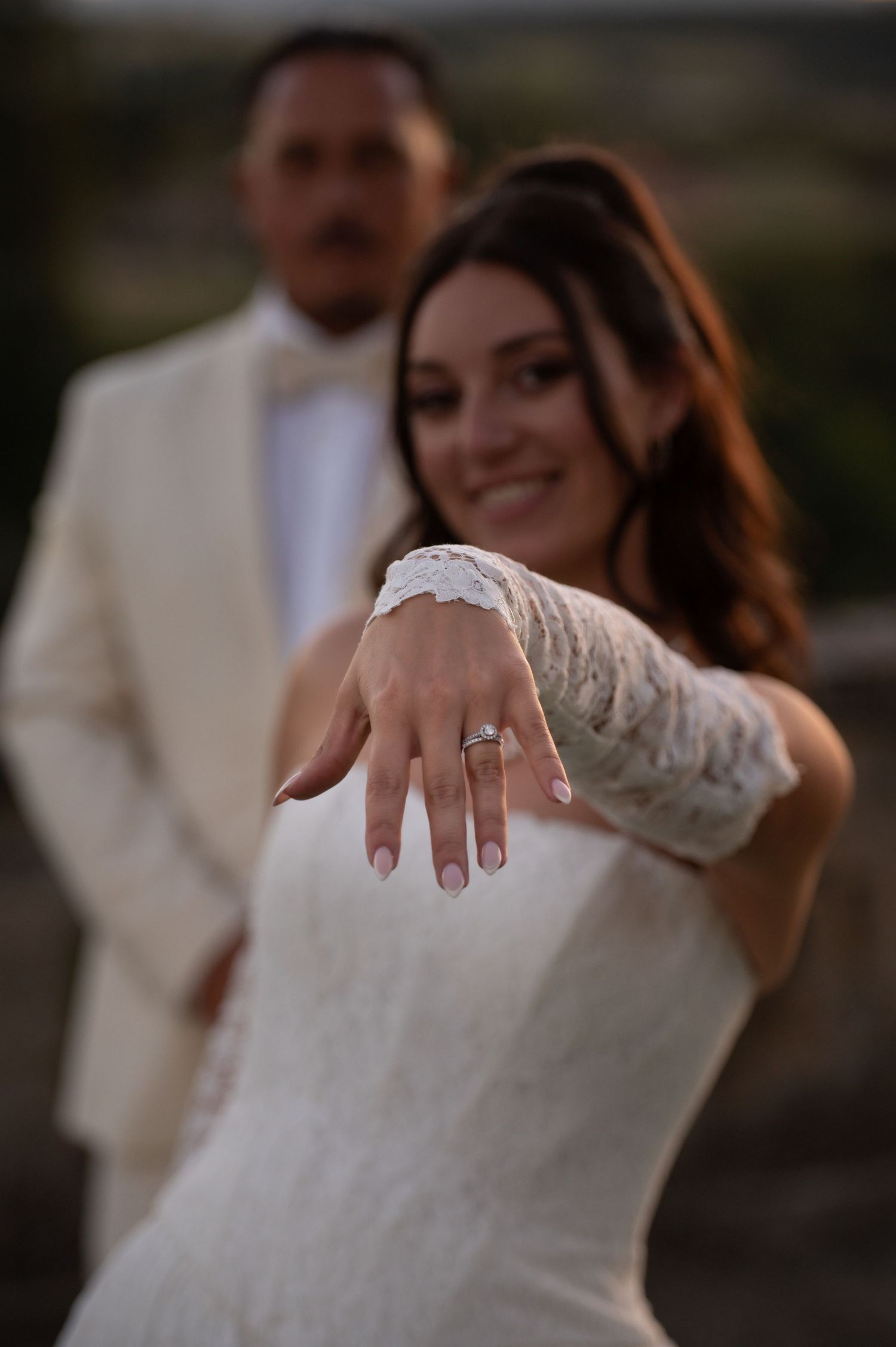 Smiling bride in white lace gown shows off diamond engagement ring, groom in cream tuxedo standing behind her.