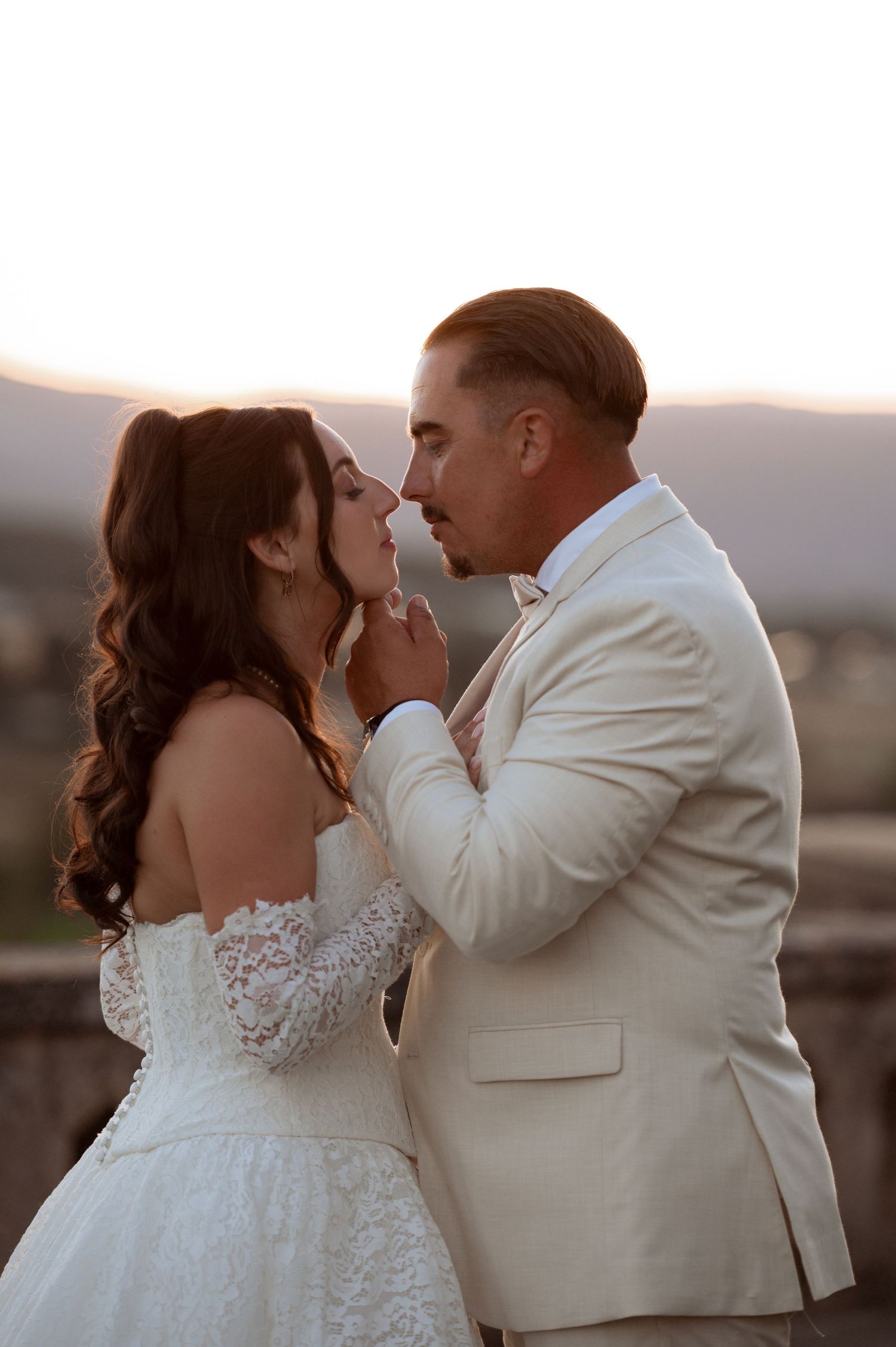 A bride and groom share a romantic moment at sunset, with the groom in a cream suit gently holding the bride's face.