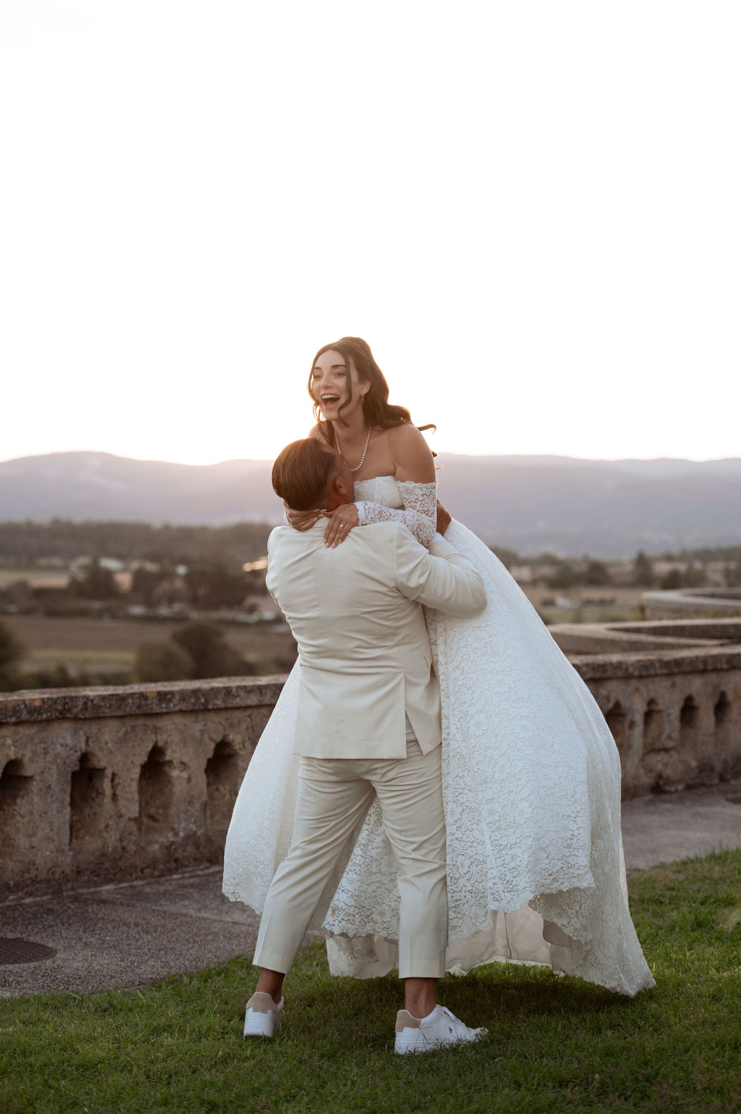 Groom lifting joyful bride in white wedding dress outdoors at sunset with scenic mountain backdrop.