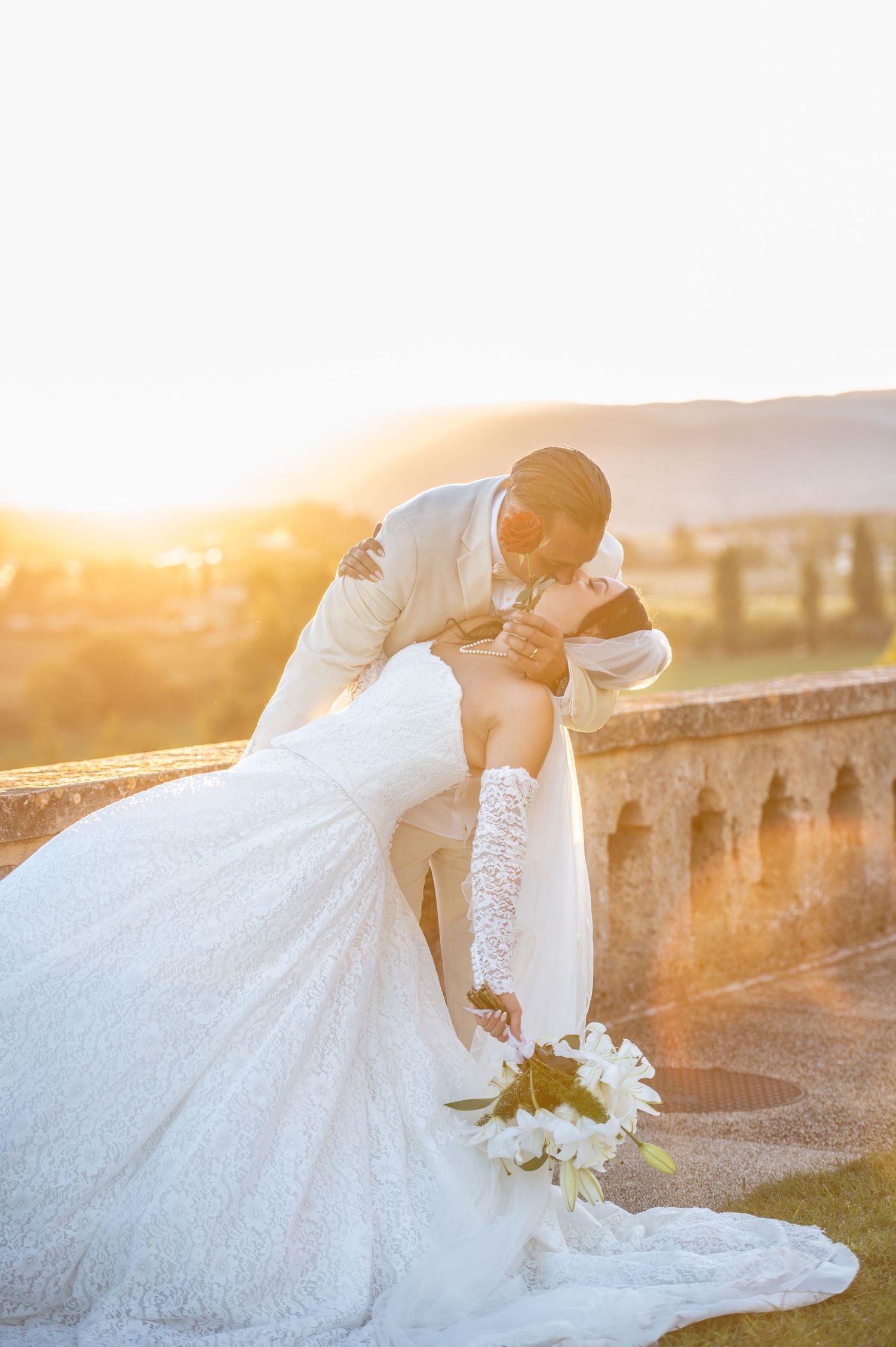 Bride and groom share a romantic dip kiss at golden hour on a stone terrace, bride holding white bouquet in lace gown.