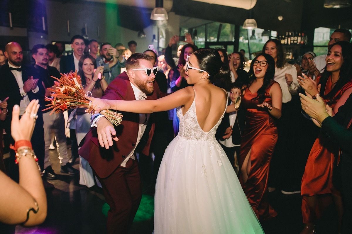 Wedding guests dance and celebrate on a crowded dance floor with colorful party lighting in the background.