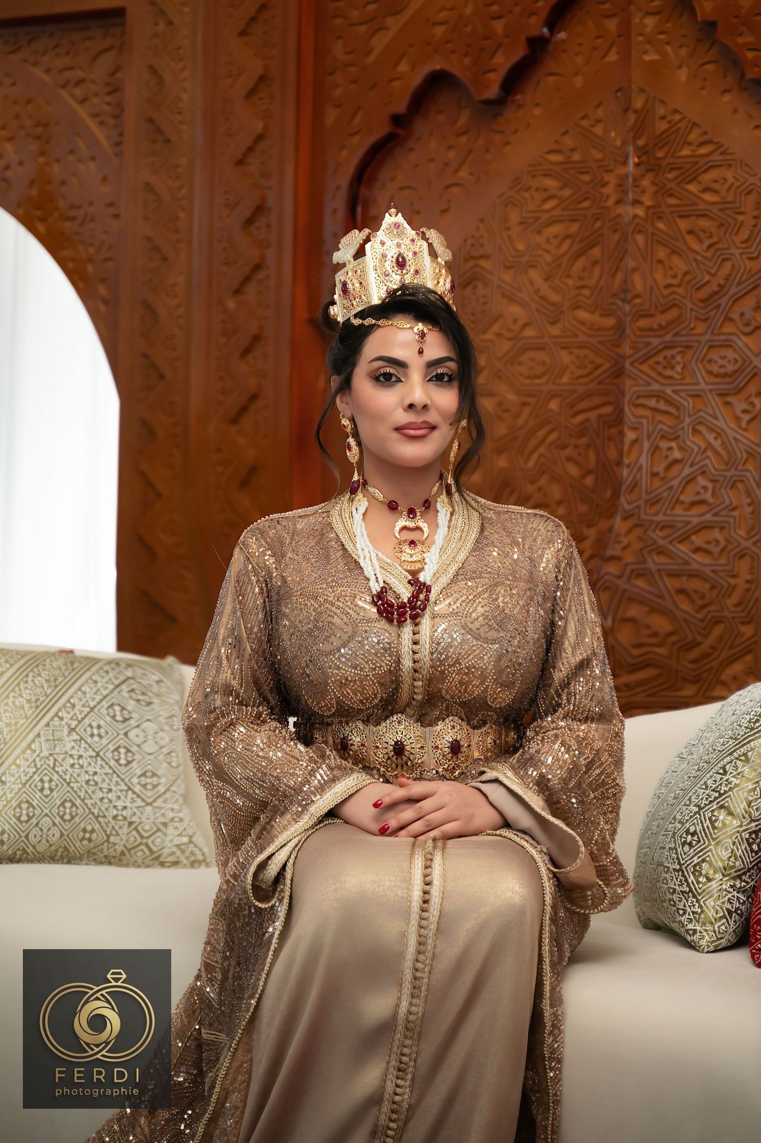 Traditional Moroccan bride wearing an elegant beige lace caftan and ornate gold crown in an ornately decorated setting.