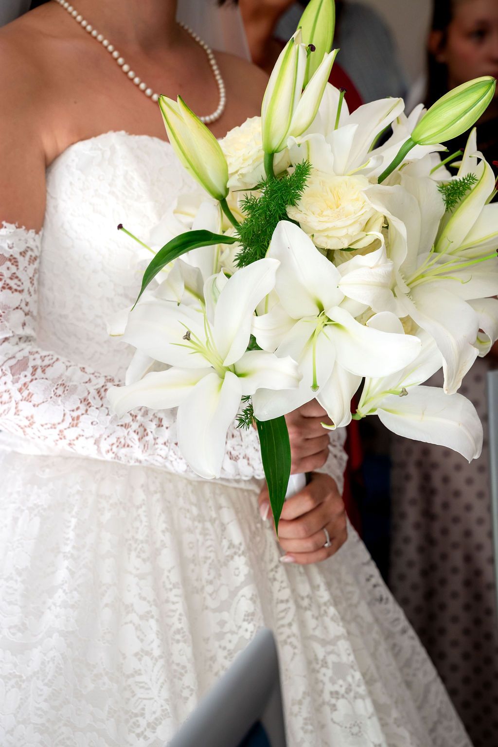 Bride in white lace gown holding elegant bouquet of white lilies and roses at wedding ceremony.