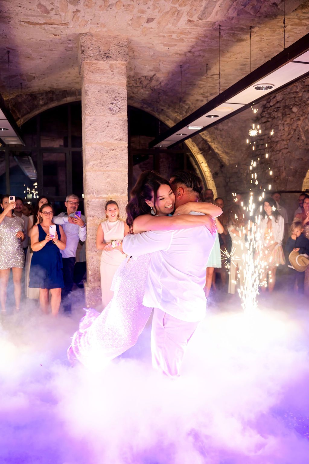 Couple shares first dance in foggy stone venue with sparkling fireworks and guests watching at a romantic wedding reception.