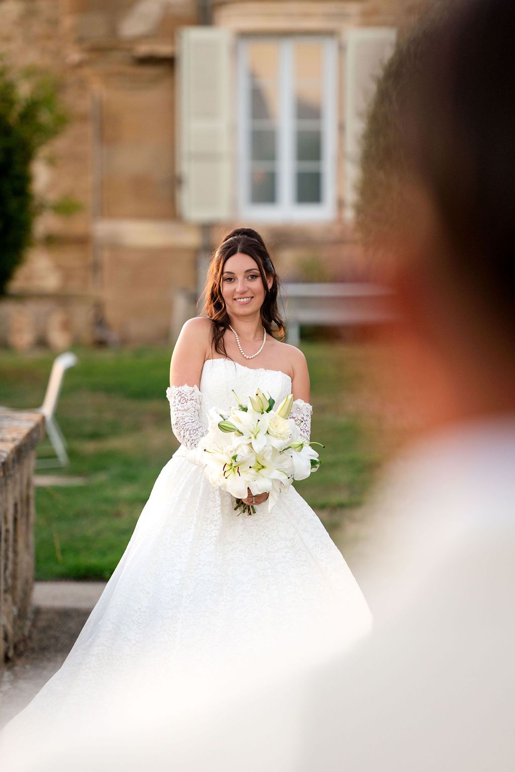 Smiling bride in white lace gown and gloves holds white lily bouquet outdoors near stone building.