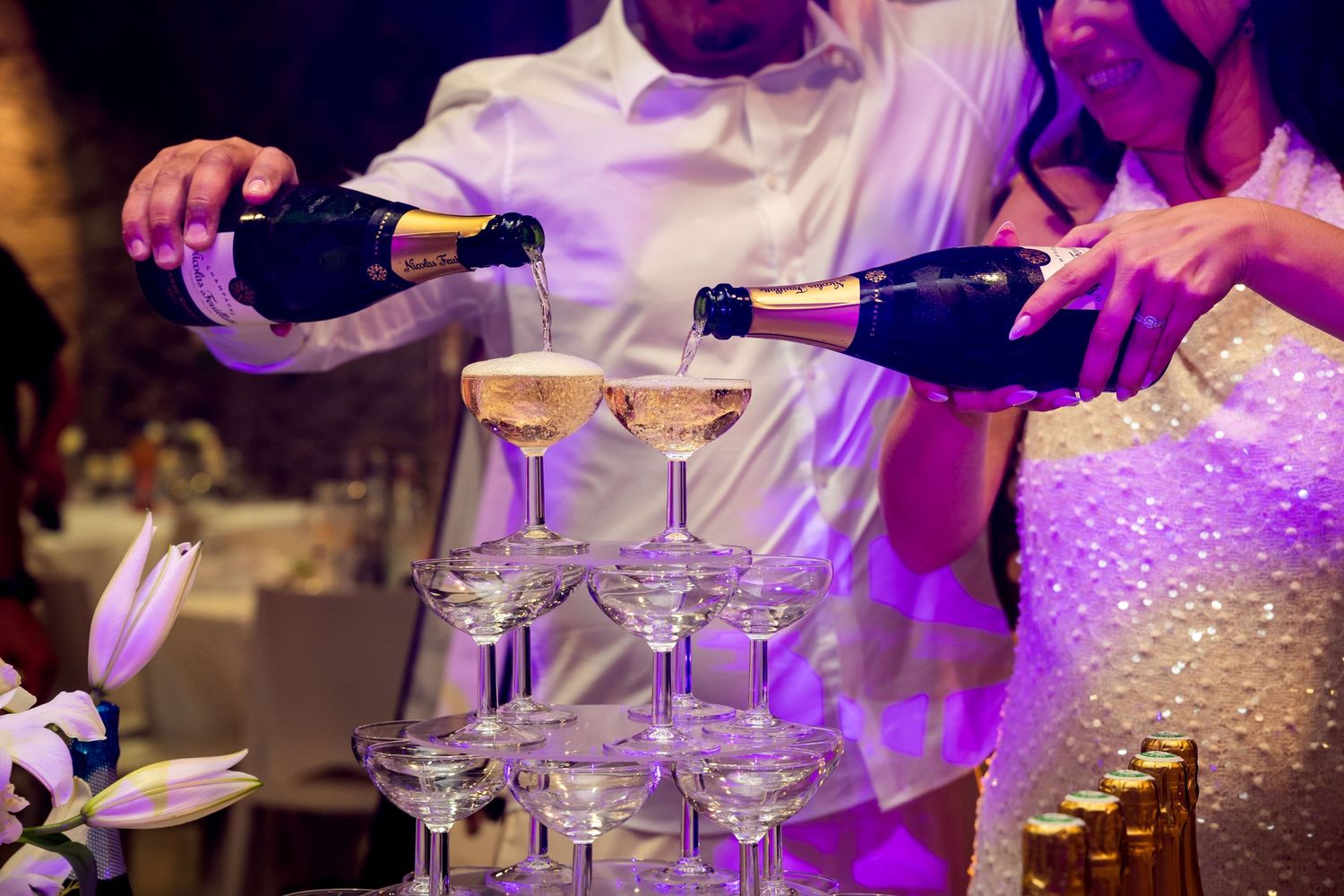 Couple pouring champagne into elegant tower of glasses at a wedding reception celebration.