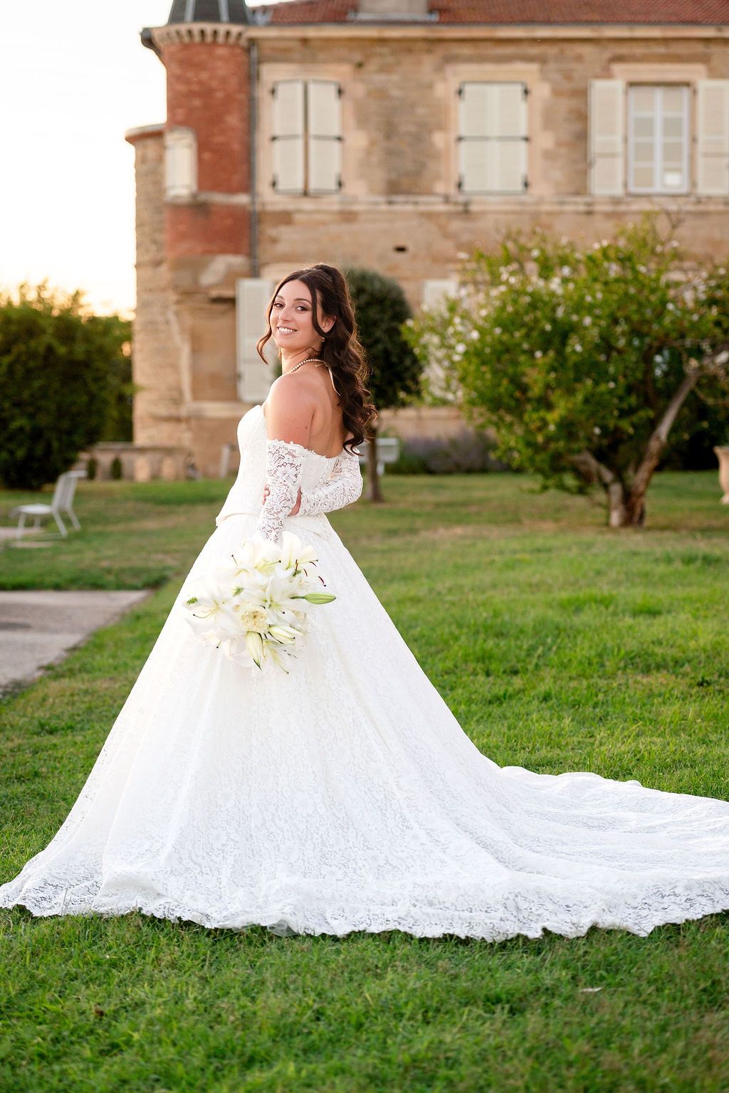 Bride in off-shoulder lace ball gown with long train holds white lily bouquet on green lawn before historic brick mansion.