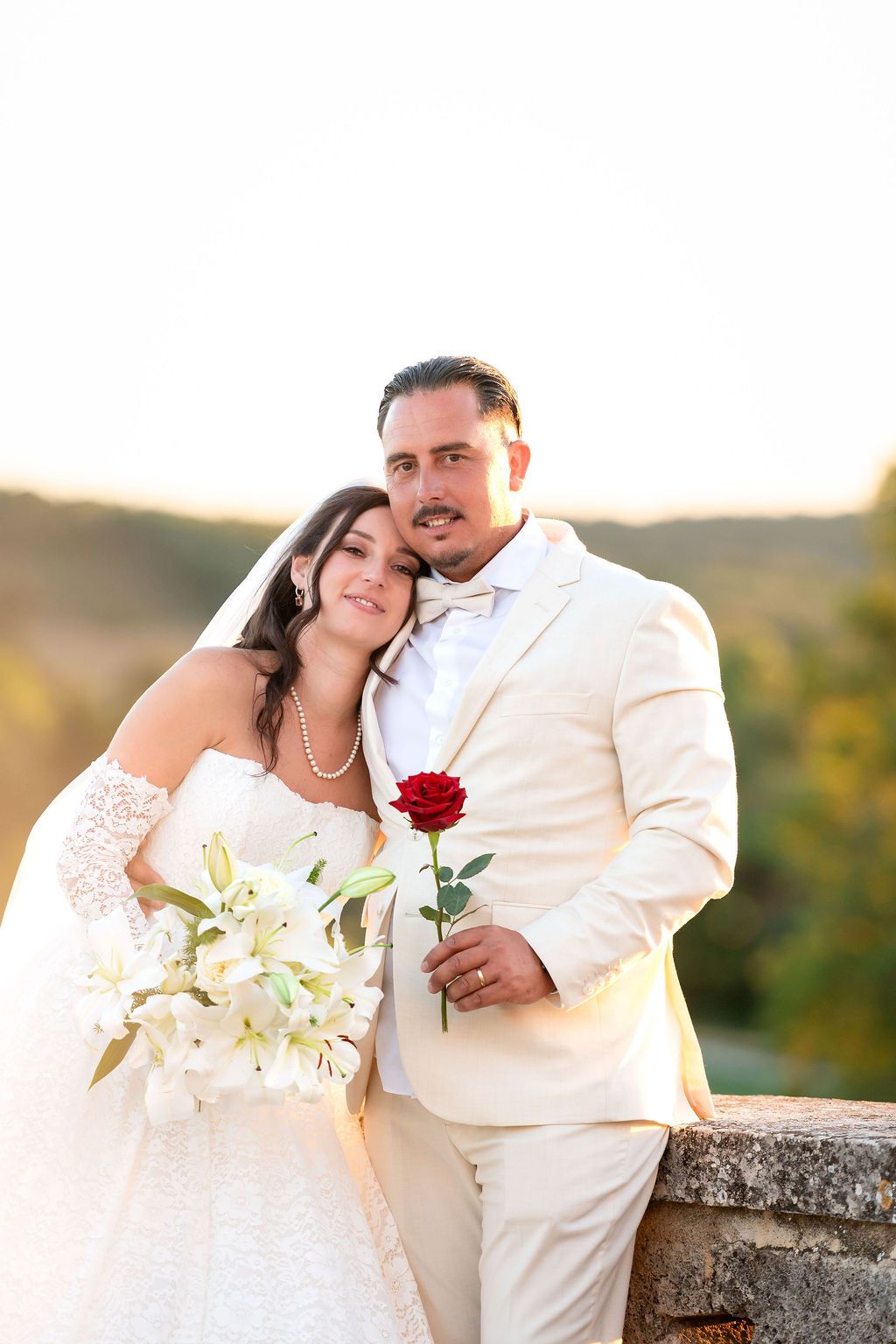 Bride and groom pose outdoors at golden hour, she holds white lilies, he holds a red rose, both in elegant wedding attire.