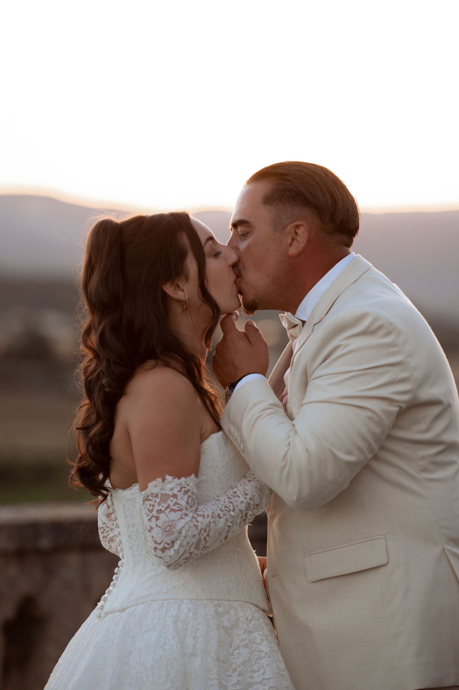 A bride and groom share a romantic kiss at sunset, she in a lace off-shoulder gown, he in a cream suit.