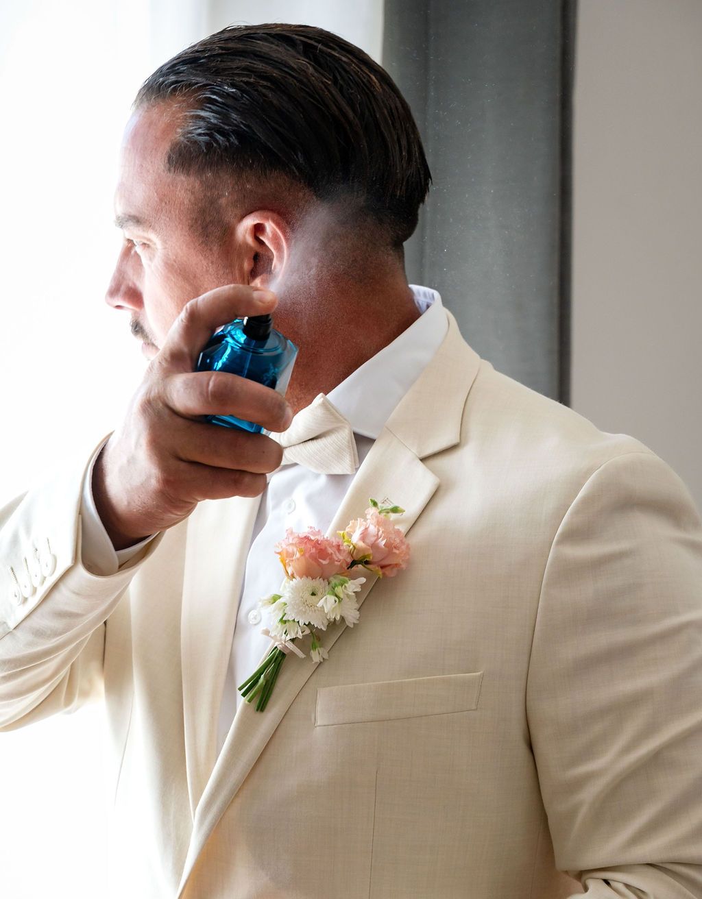 Groom in beige suit with floral boutonniere applying cologne before wedding ceremony.