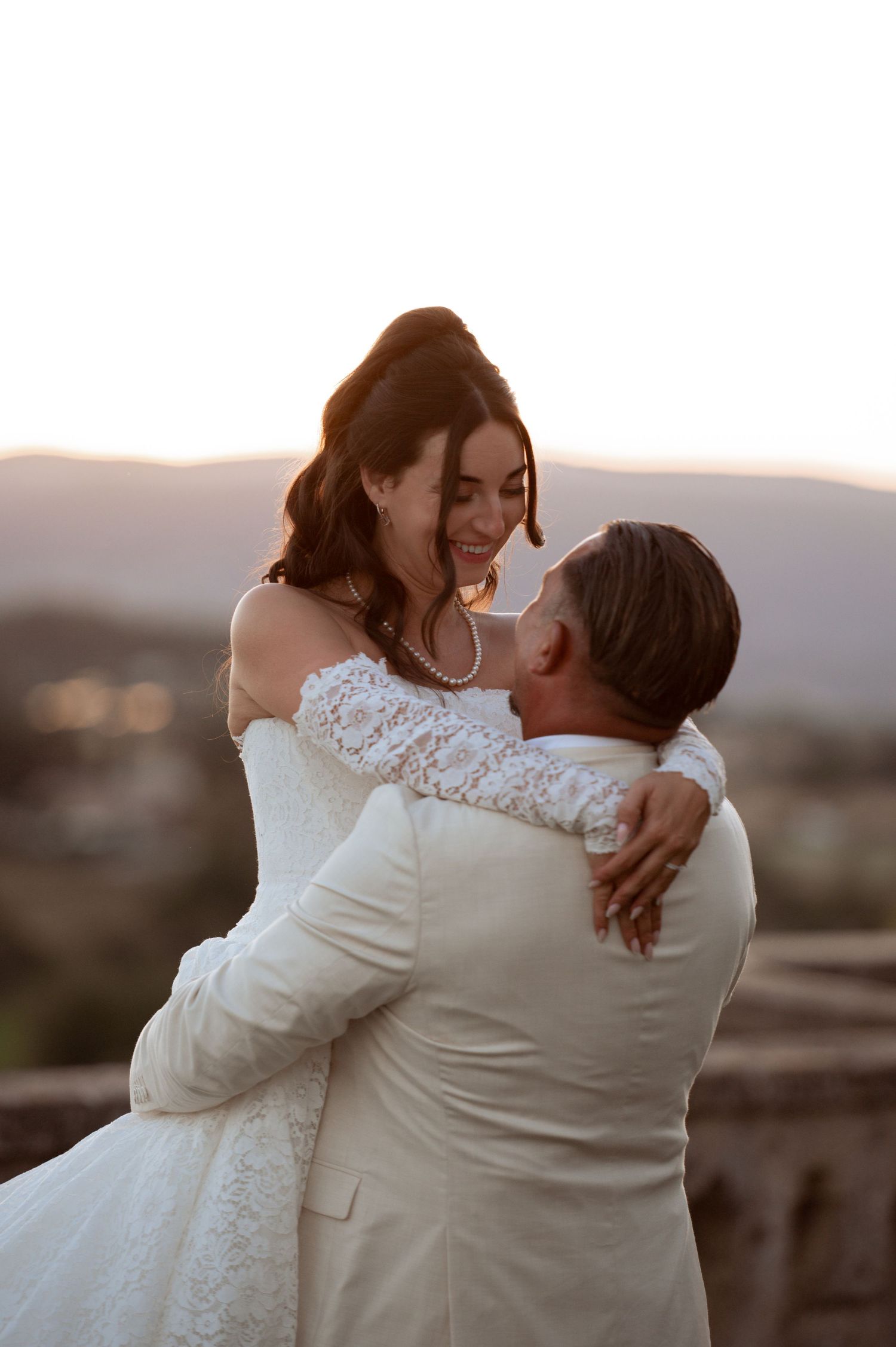 A groom in a white suit lifts his smiling bride in a lace off-shoulder gown during a golden hour wedding portrait.