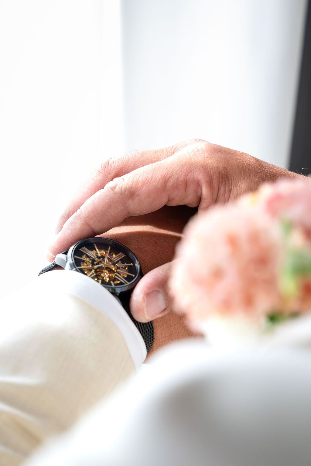 Close-up of a man's wrist wearing a luxury skeleton mechanical watch with gold details, beside pink flowers.