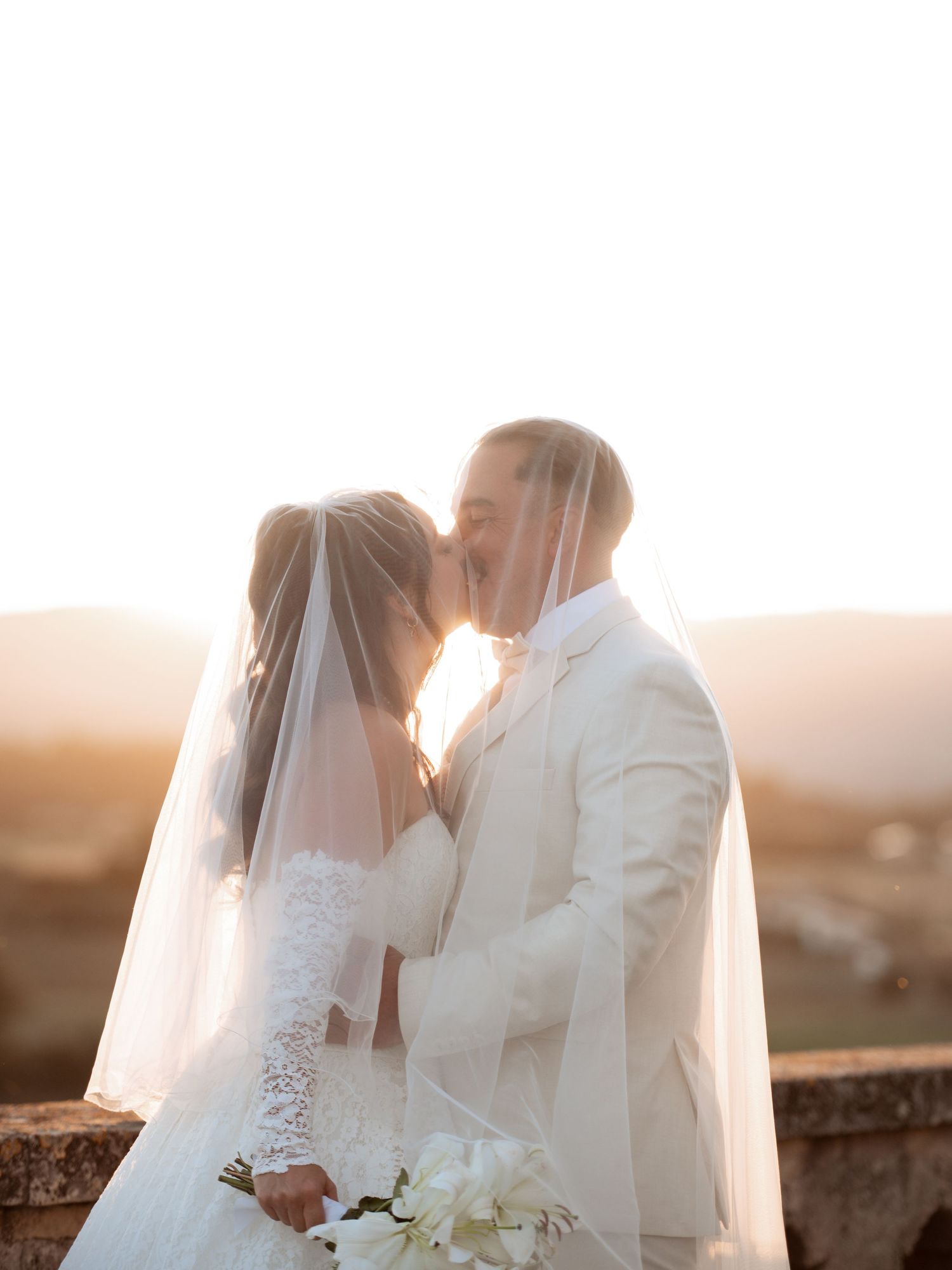 Bride and groom sharing a romantic sunset kiss, bride in lace gown with veil, groom in white suit, holding white bouquet.