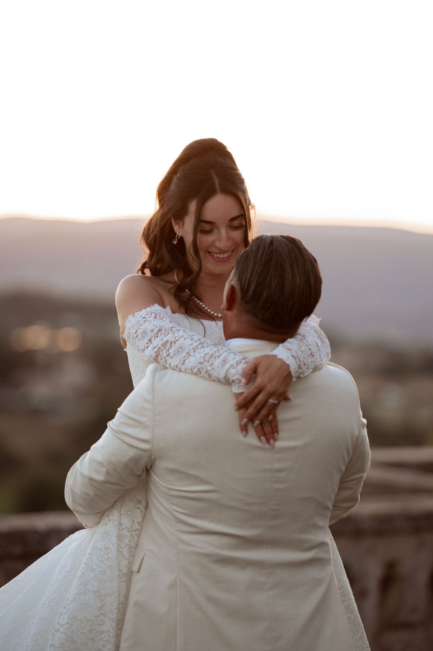 Bride in lace off-shoulder gown embraces groom in white suit at golden hour outdoor wedding ceremony.