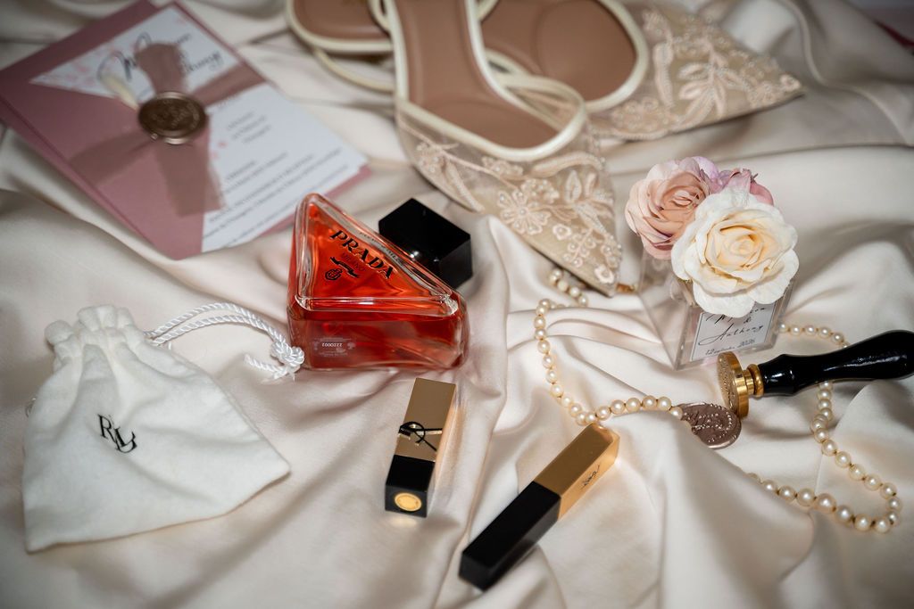 Bridal flat lay with perfume, lipstick, pearl necklace, heels, pink rose, and wedding invitation on white satin.