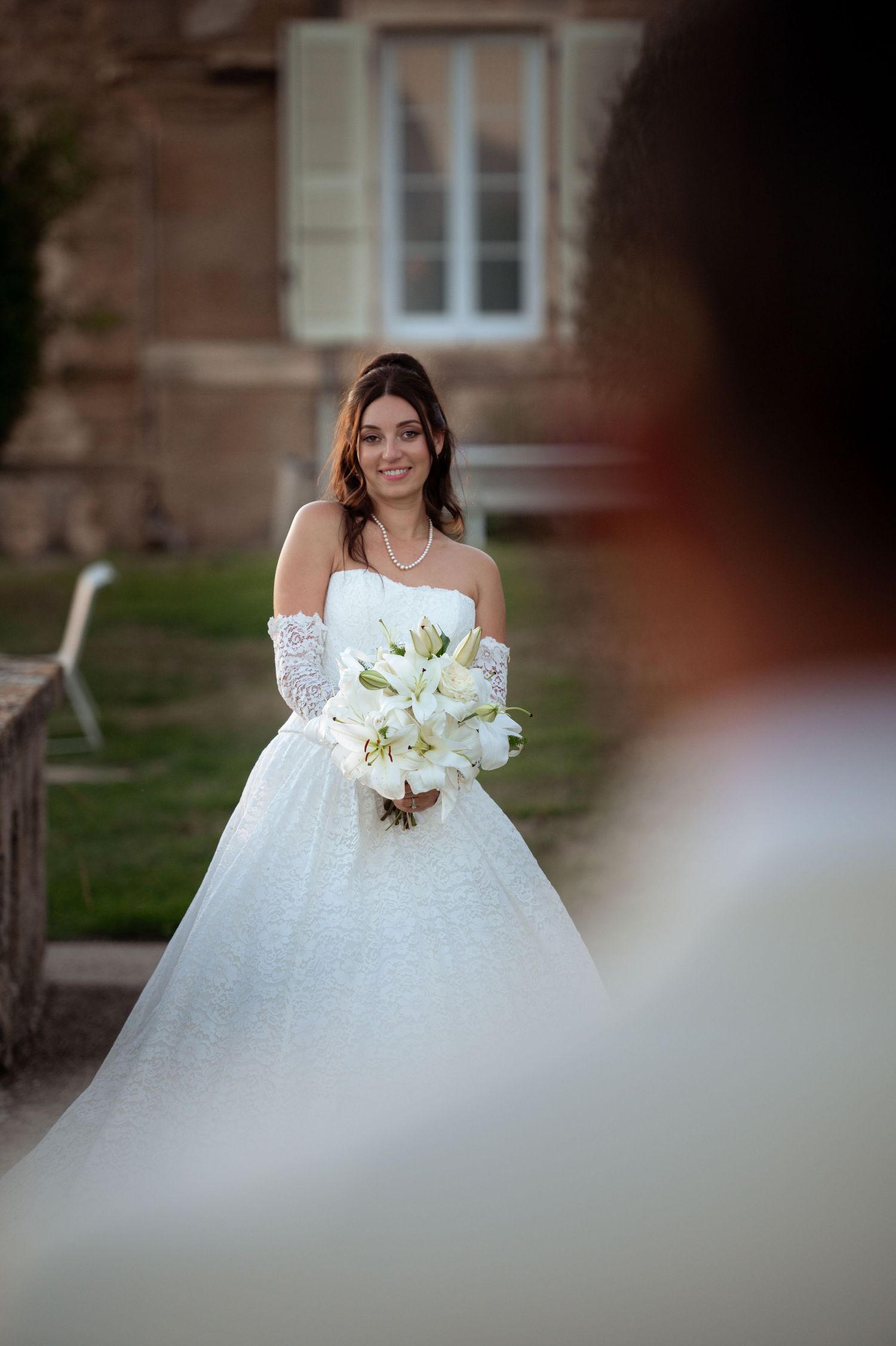 Smiling bride in white ball gown holding white lily bouquet, wearing lace gloves and pearl necklace outdoors.