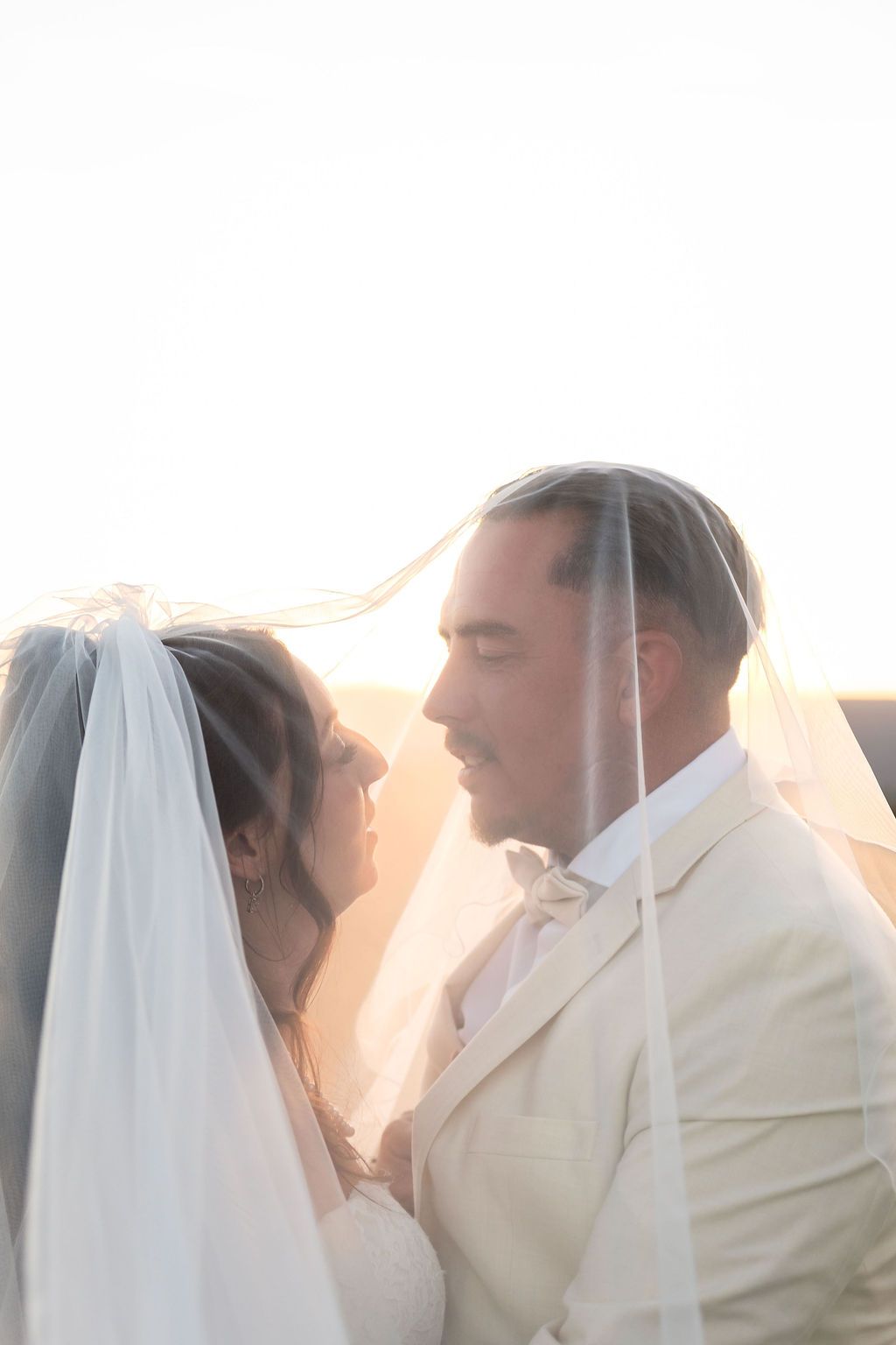 Bride and groom share romantic moment under flowing veil at golden hour sunset wedding.