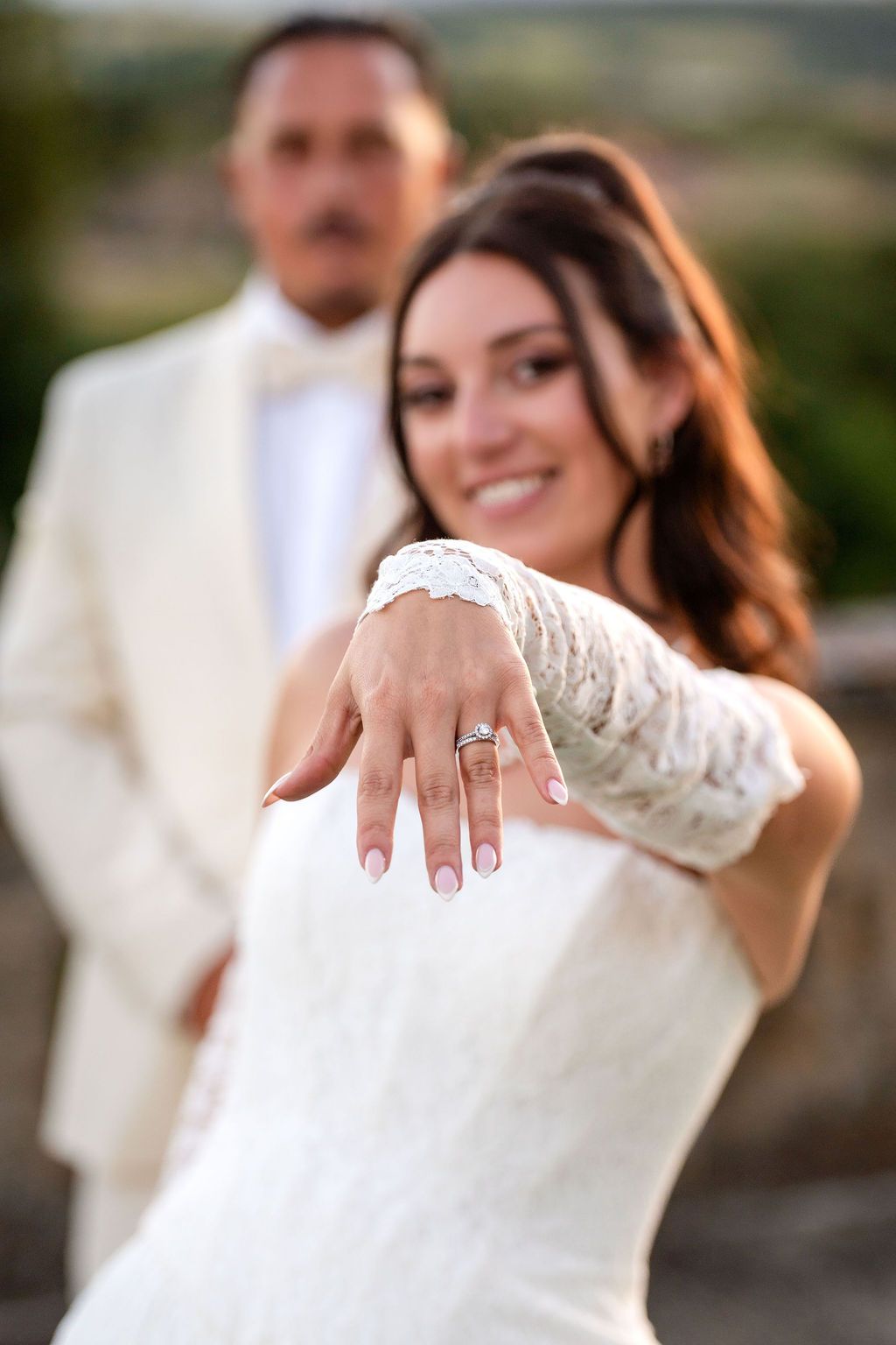 Smiling bride in white lace dress shows off wedding ring while groom in cream suit stands behind her outdoors.