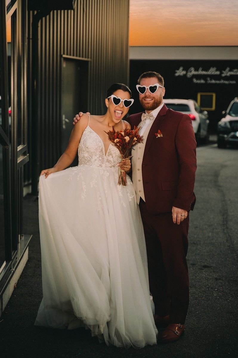 A couple wearing sunglasses pose together at sunset, with the bride in a flowing white gown and groom in burgundy suit.