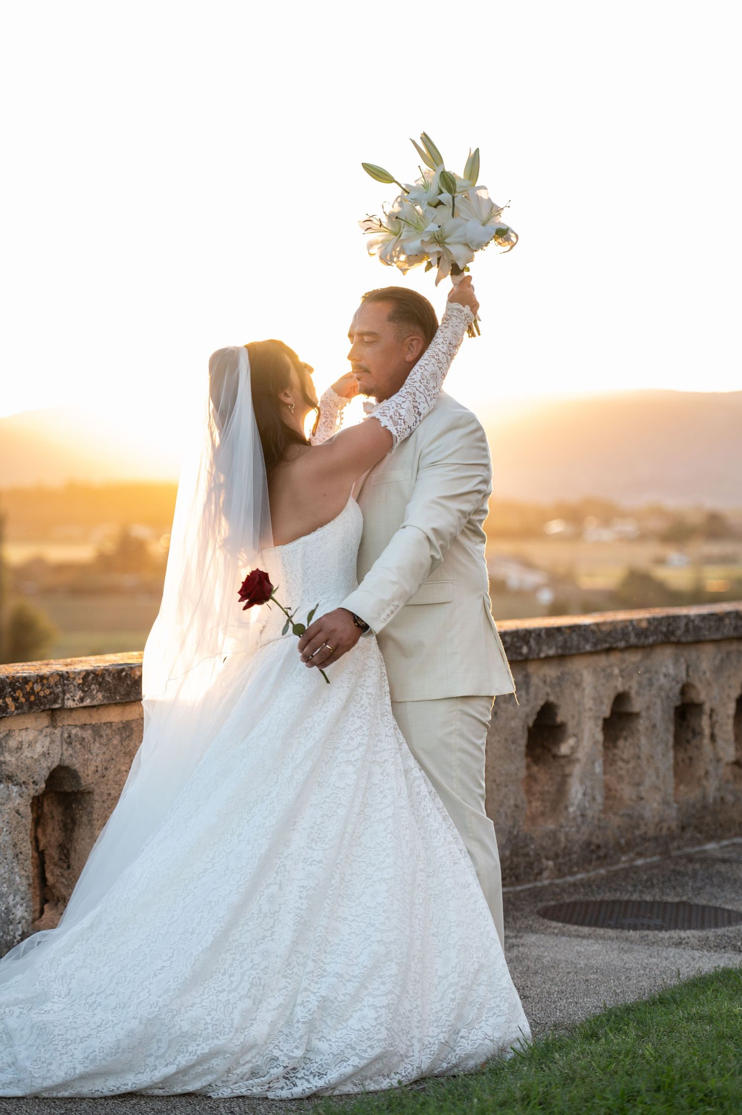 Bride and groom share a romantic sunset kiss on a stone balcony, she holds a white bouquet aloft while he holds a red rose.